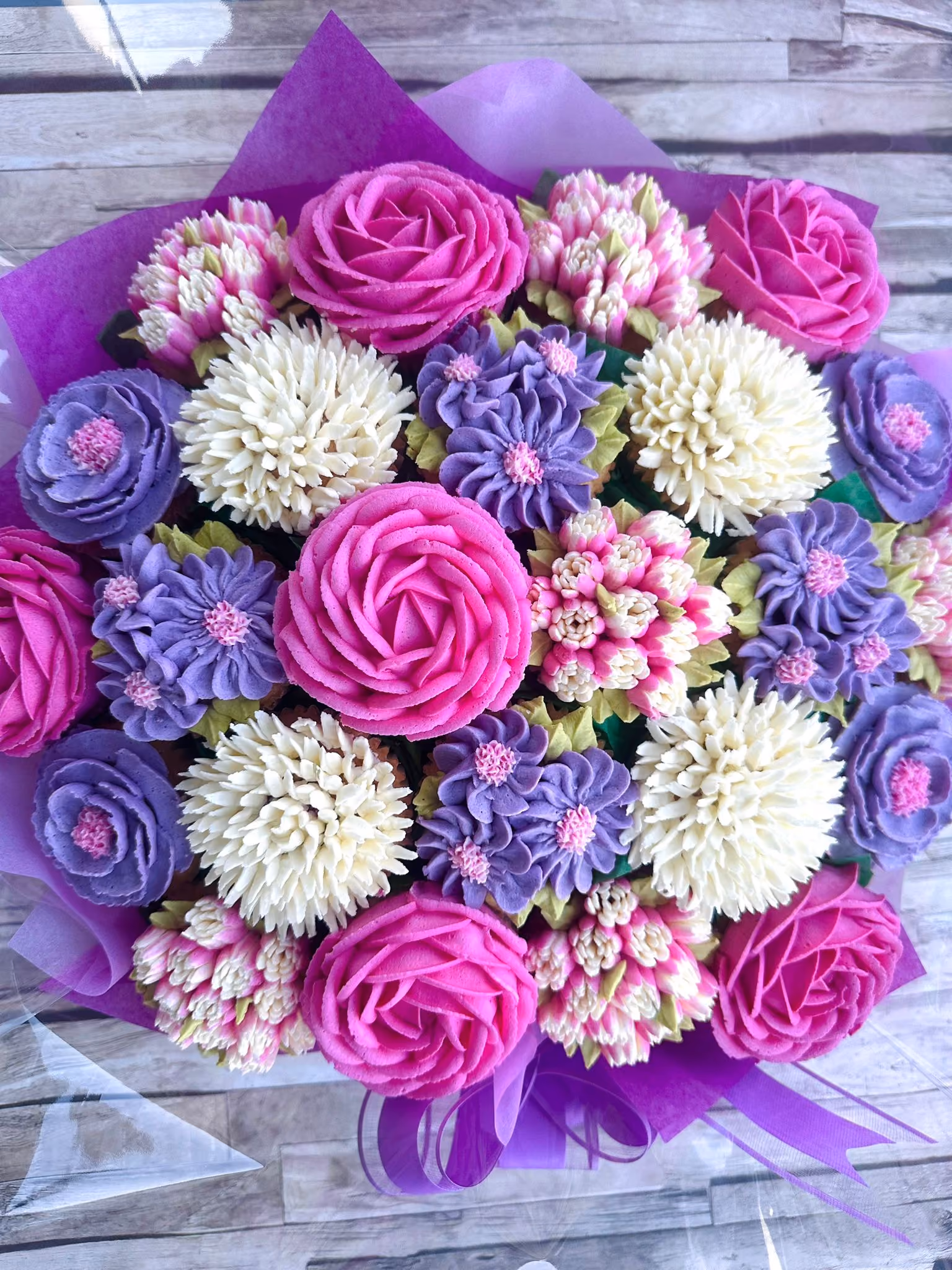 Colorful bouquet of pink, purple, and white flowers intricately made from piped frosting, arranged with purple wrapping and ribbon on a wooden surface.