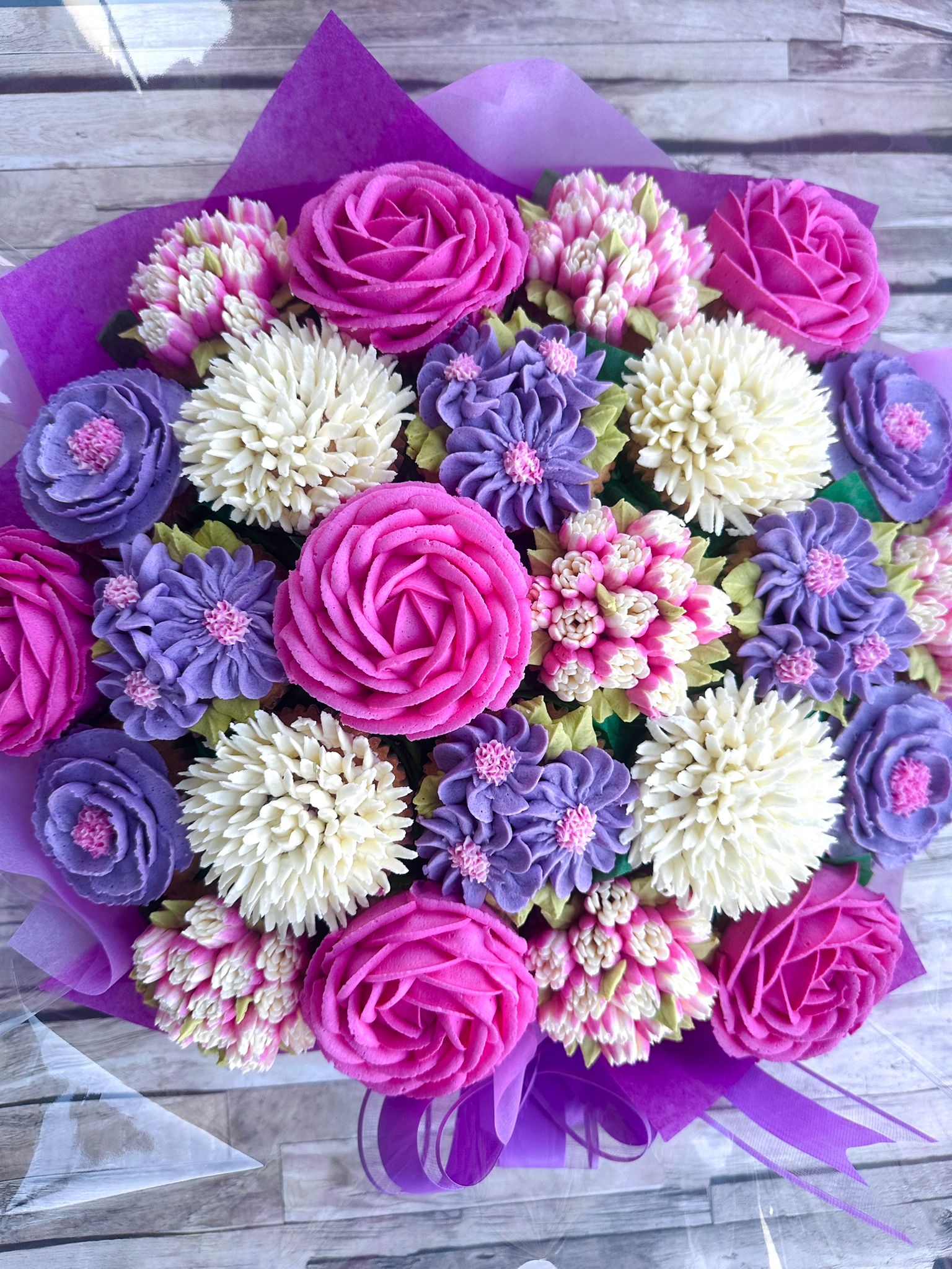 Colorful bouquet of pink, purple, and white flowers intricately made from piped frosting, arranged with purple wrapping and ribbon on a wooden surface.