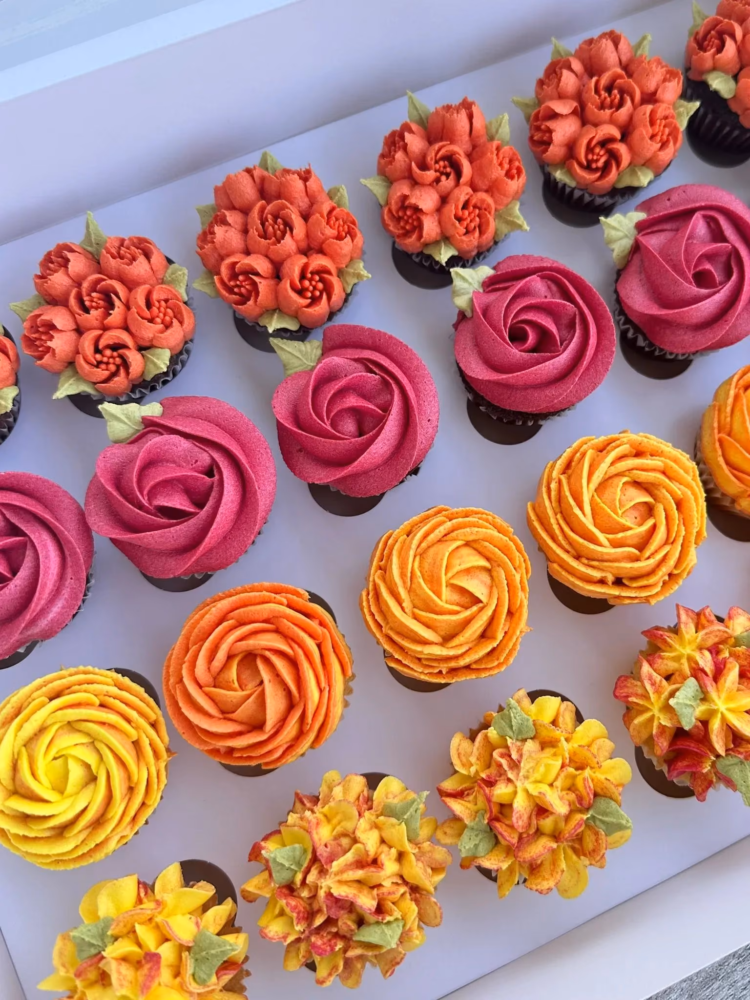 Box of colorful cupcakes decorated with orange, pink, yellow, and red floral buttercream designs.