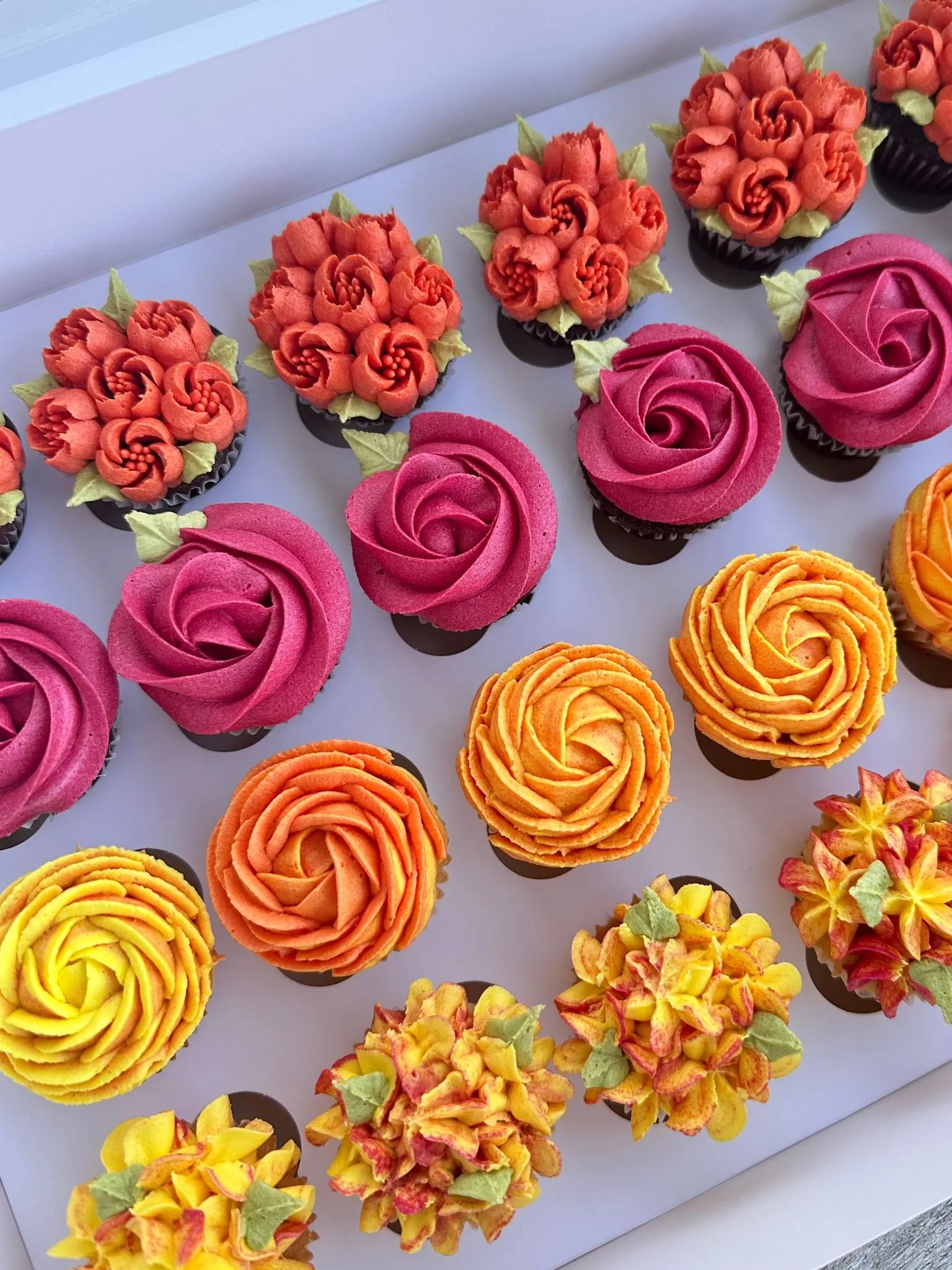 Box of colorful cupcakes decorated with orange, pink, yellow, and red floral buttercream designs.