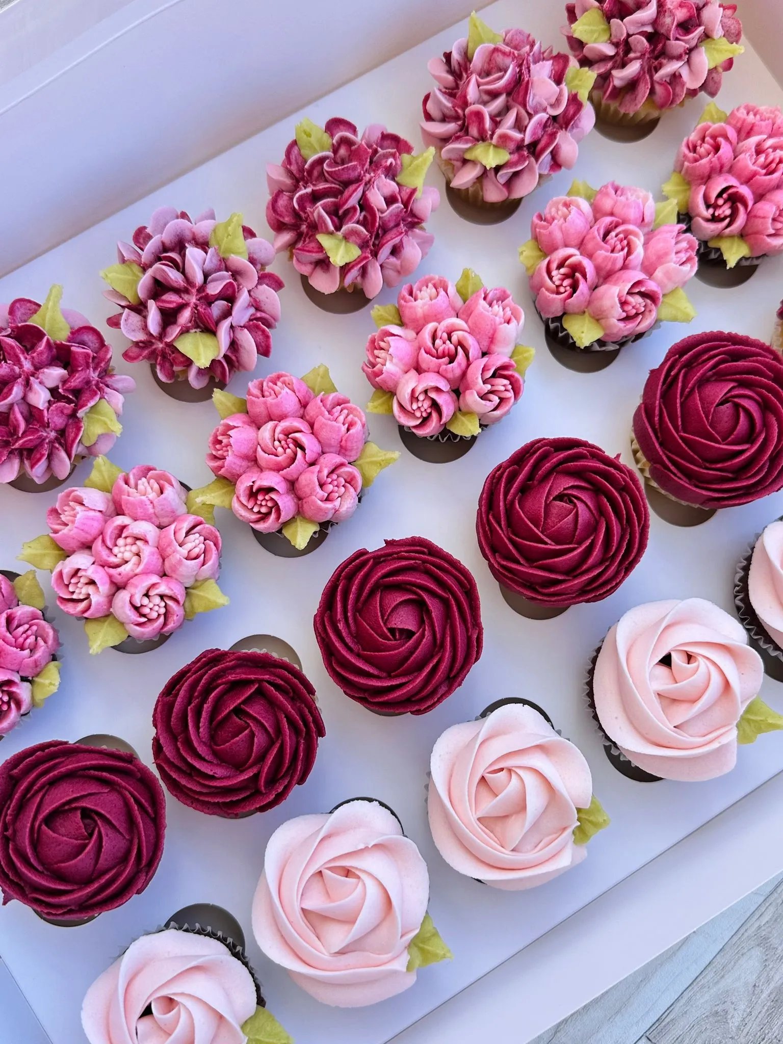 Box of assorted cupcakes decorated with pink, burgundy, and mauve buttercream flowers and green leaves.