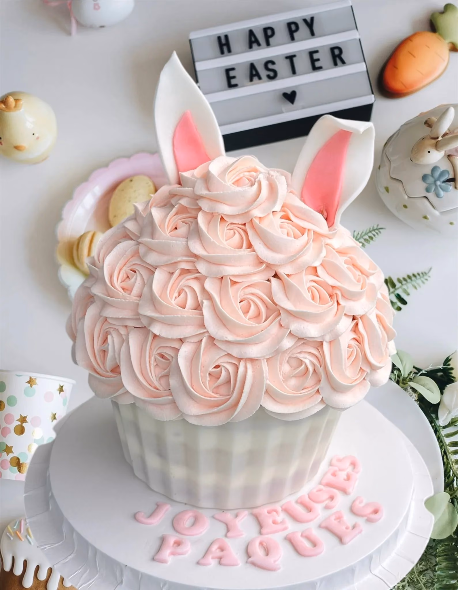 Large cupcake decorated with peach rose-shaped frosting and pearl-like accents inside a clear cylindrical gift box with a white lid and ribbon, surrounded by purple flowers.