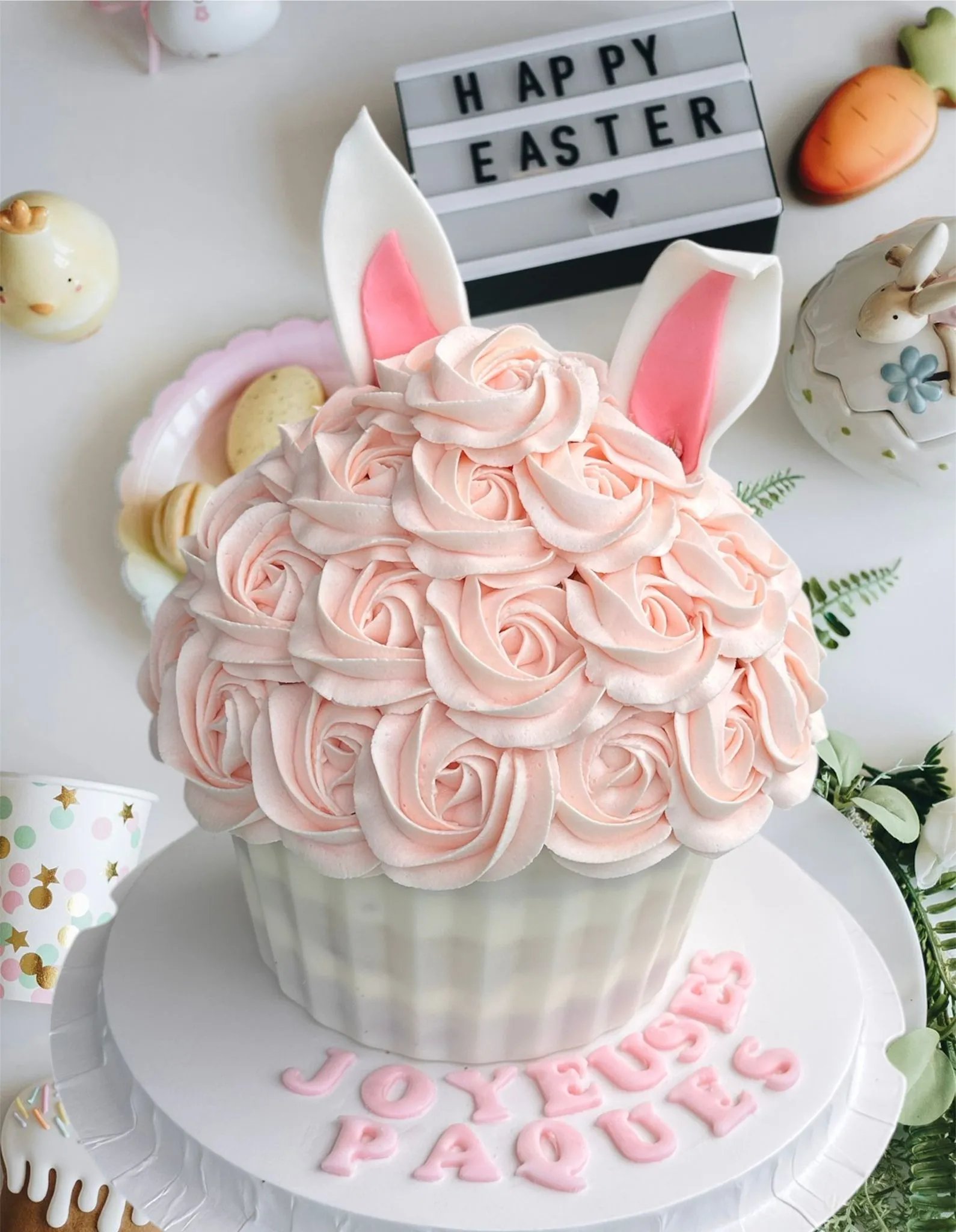 Large cupcake decorated with peach rose-shaped frosting and pearl-like accents inside a clear cylindrical gift box with a white lid and ribbon, surrounded by purple flowers.
