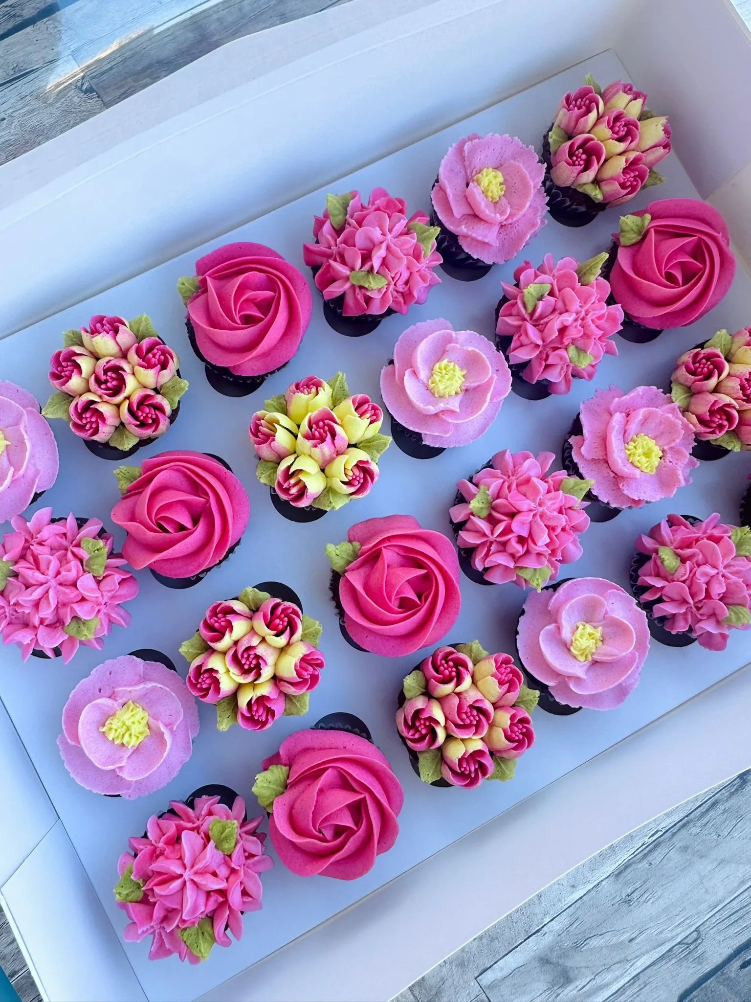 Box of twelve cupcakes decorated with intricate flower designs in pink, yellow, and red frosting with green leaves.