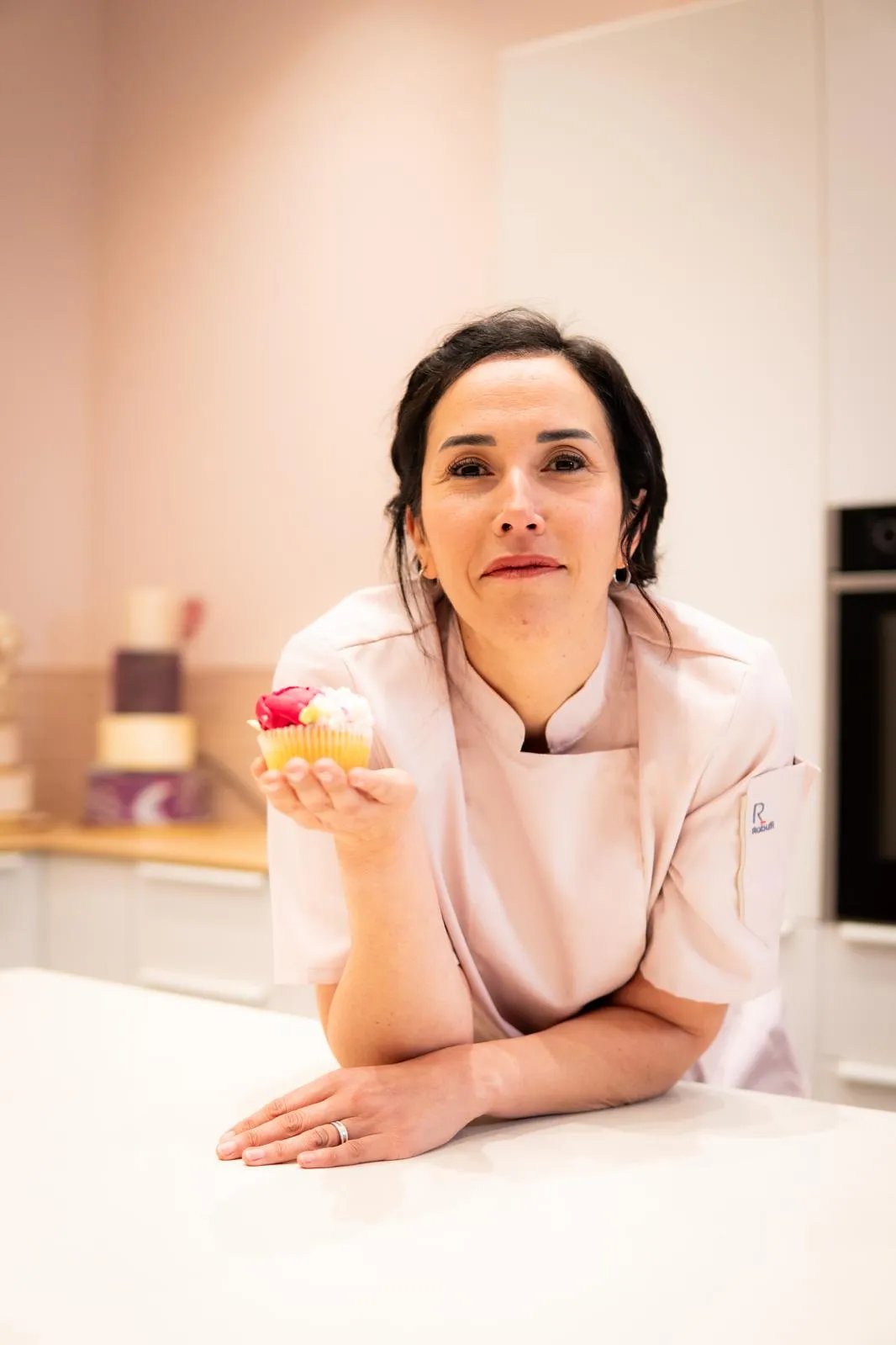 Female chef in a white uniform leaning on a kitchen counter holding a decorated cupcake with pink and white frosting.