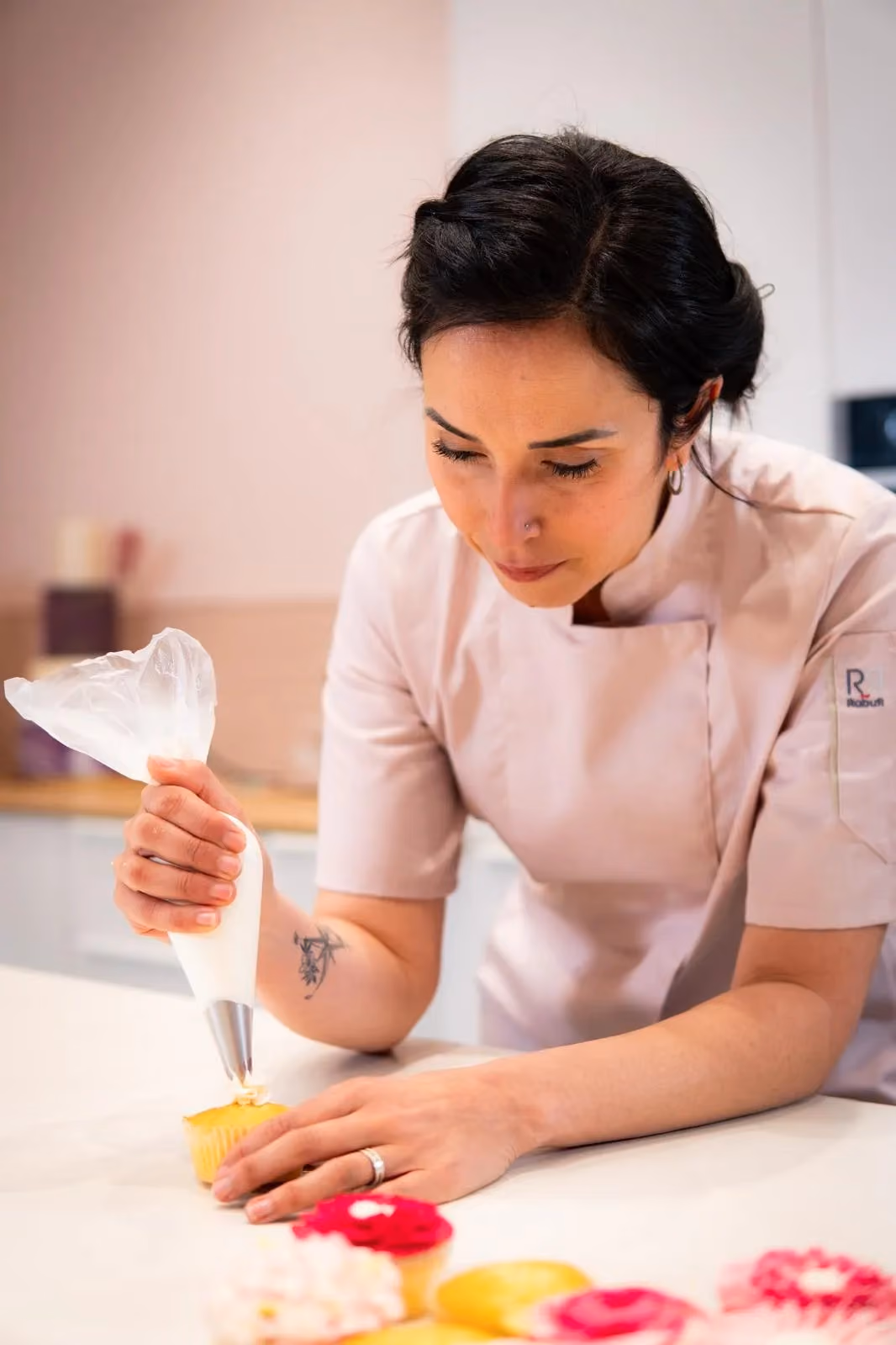 Female chef in light pink uniform decorating a yellow cupcake with white icing using a piping bag.