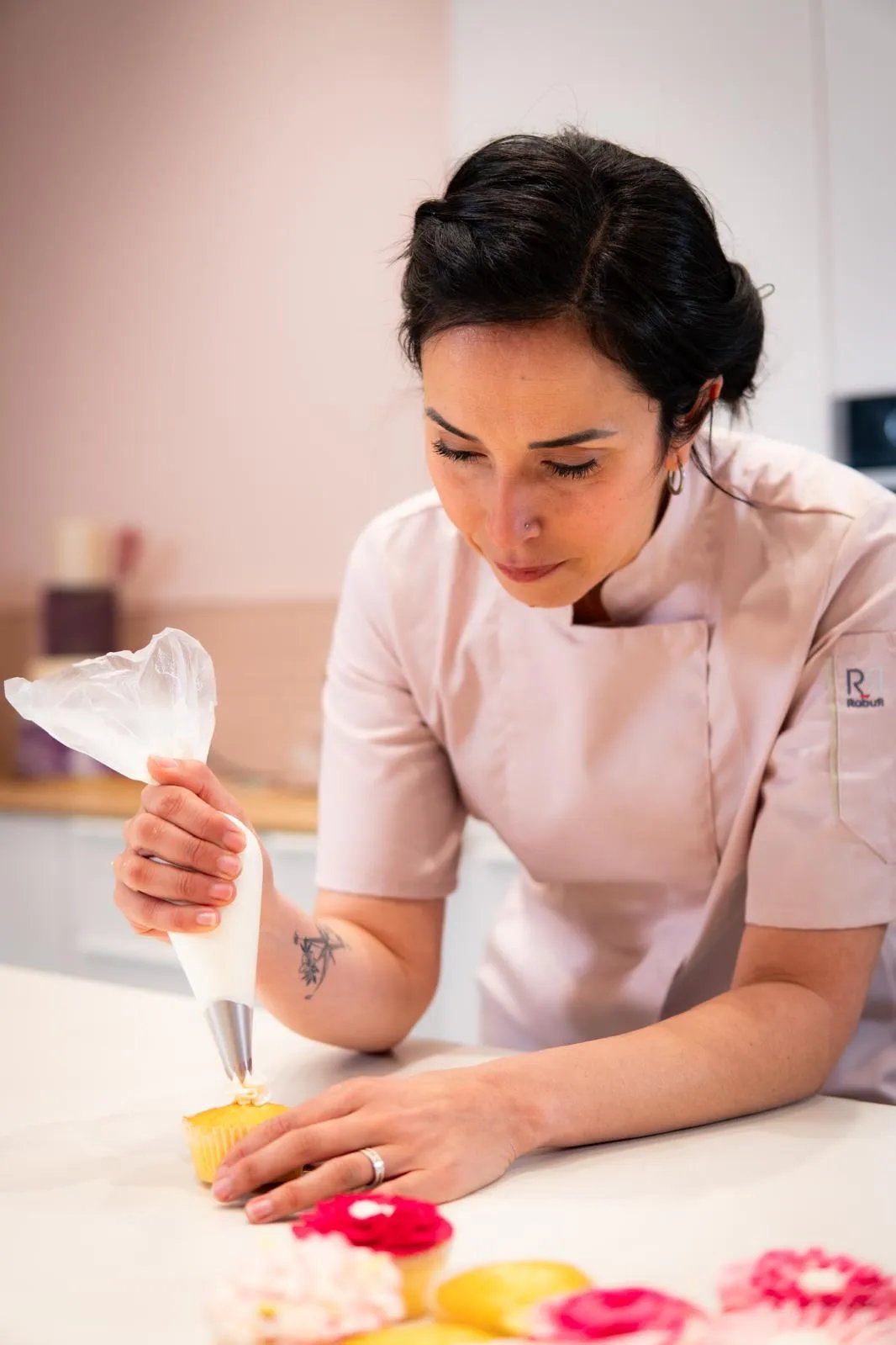Female chef in light pink uniform decorating a yellow cupcake with white icing using a piping bag.