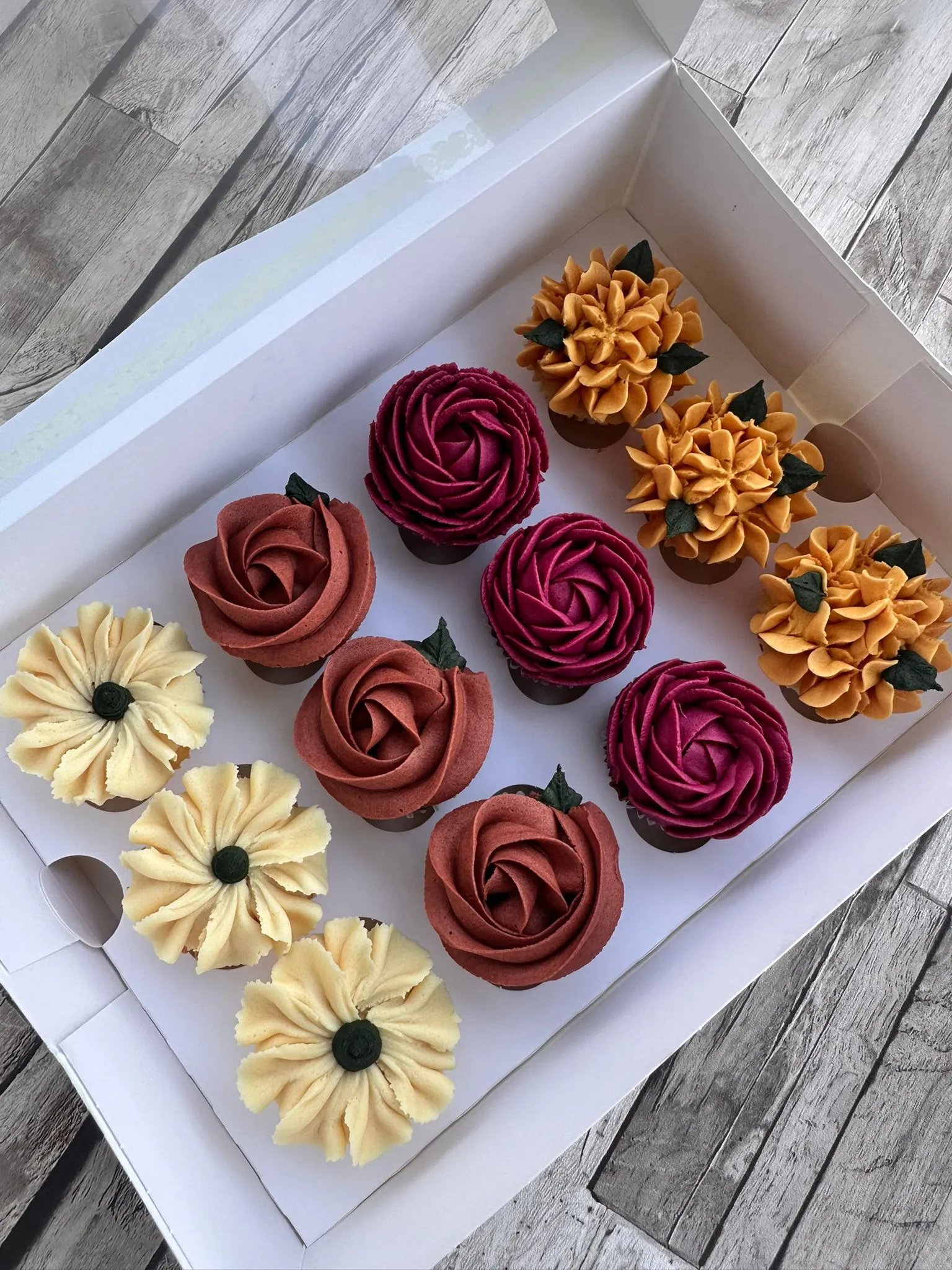 Box of twelve cupcakes decorated with cream frosting in red, yellow, orange, and purple floral designs on a gray wooden surface.
