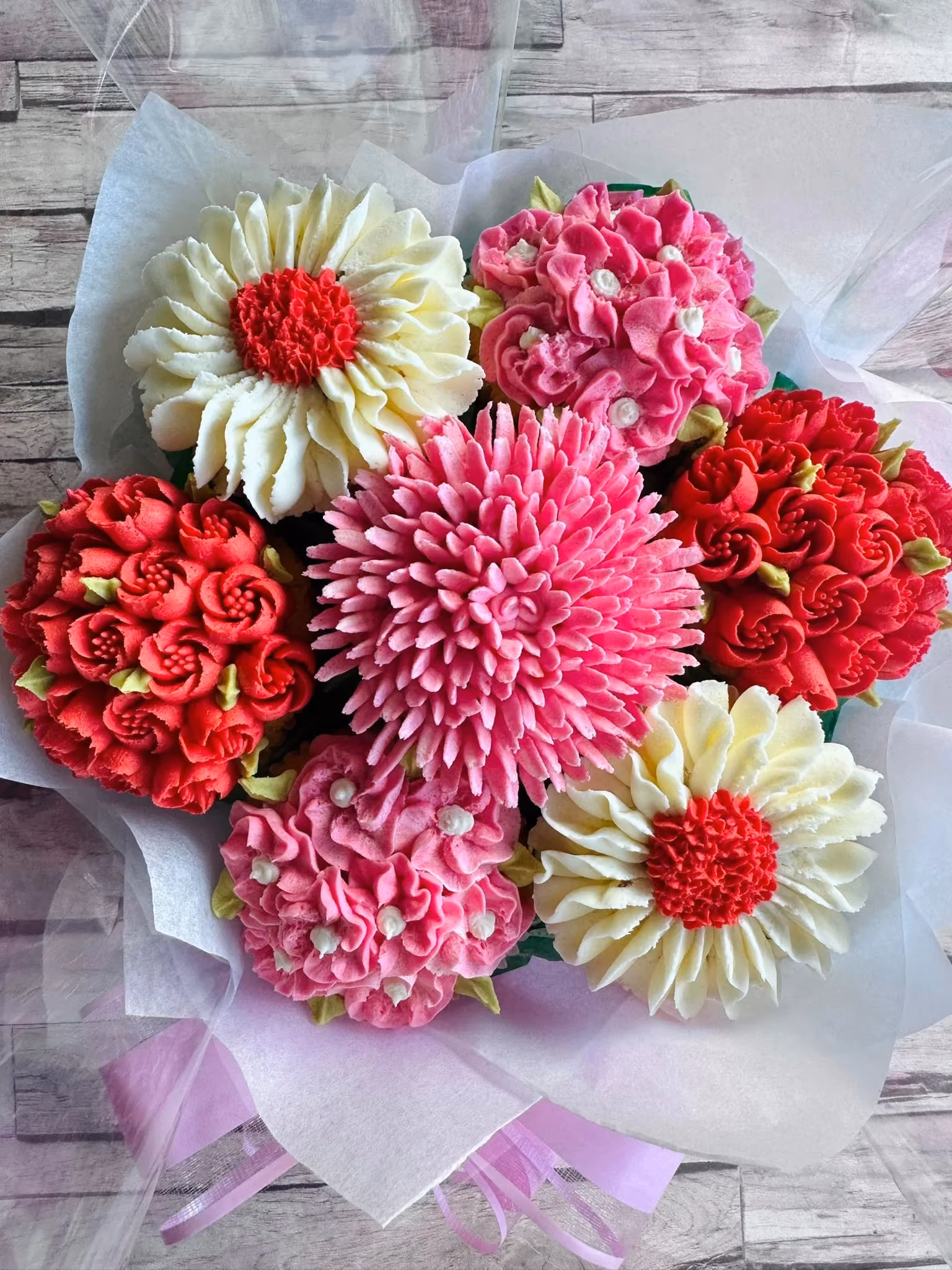 Bouquet of intricately decorated cupcakes with floral frosting in red, pink, and white colors on a wooden surface.