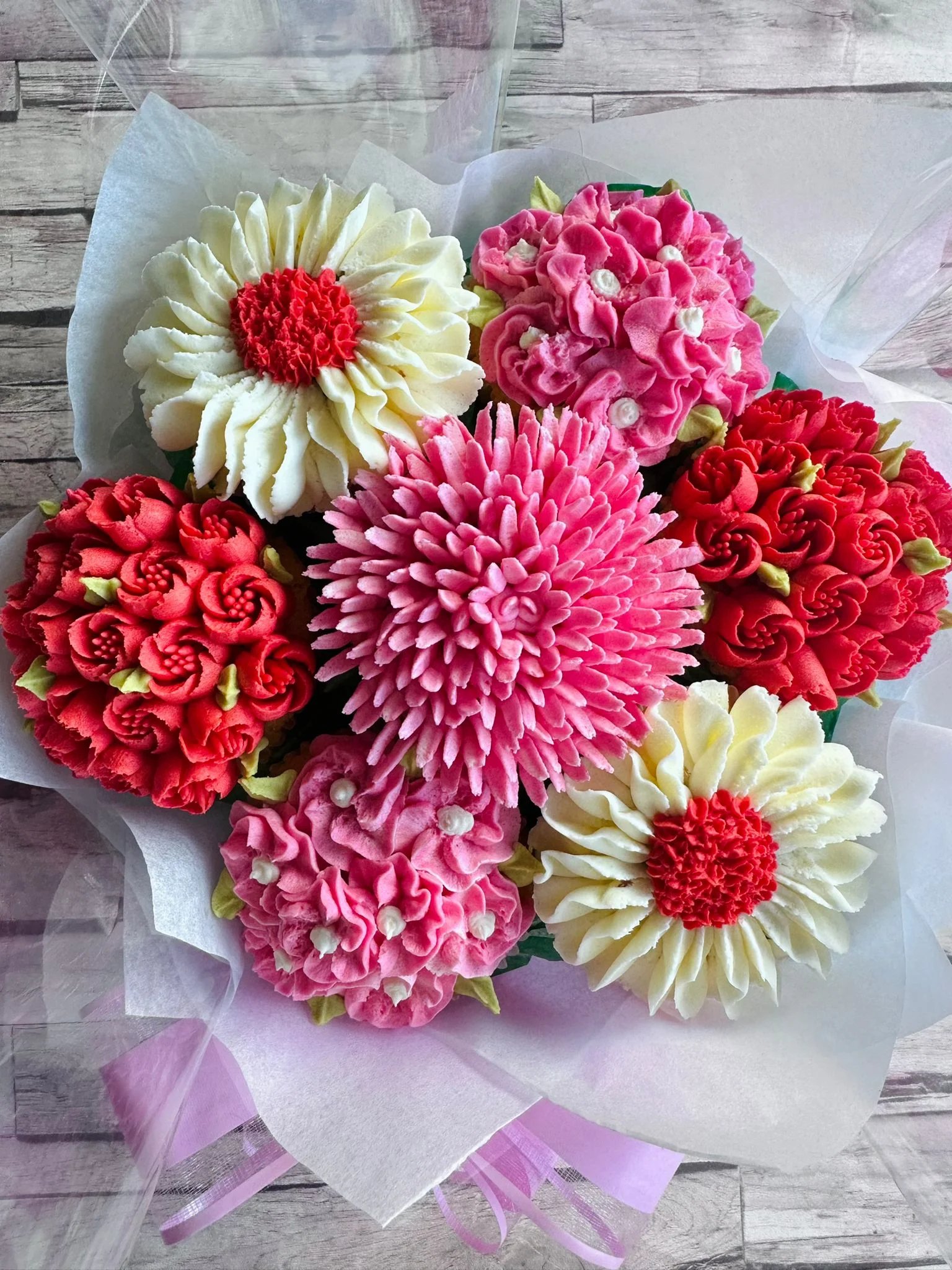 Bouquet of intricately decorated cupcakes with floral frosting in red, pink, and white colors on a wooden surface.