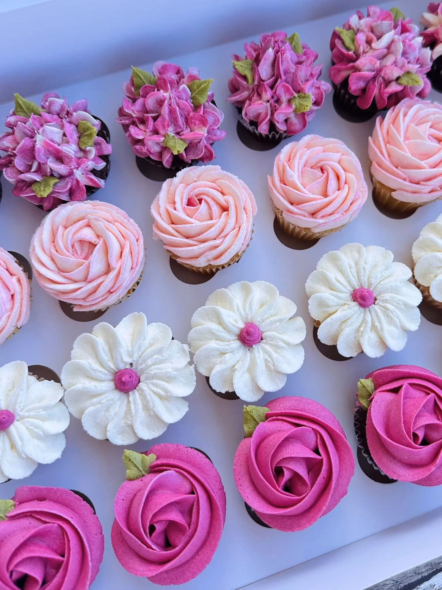 Box of assorted cupcakes decorated with pink, white, and purple flower-themed frosting.