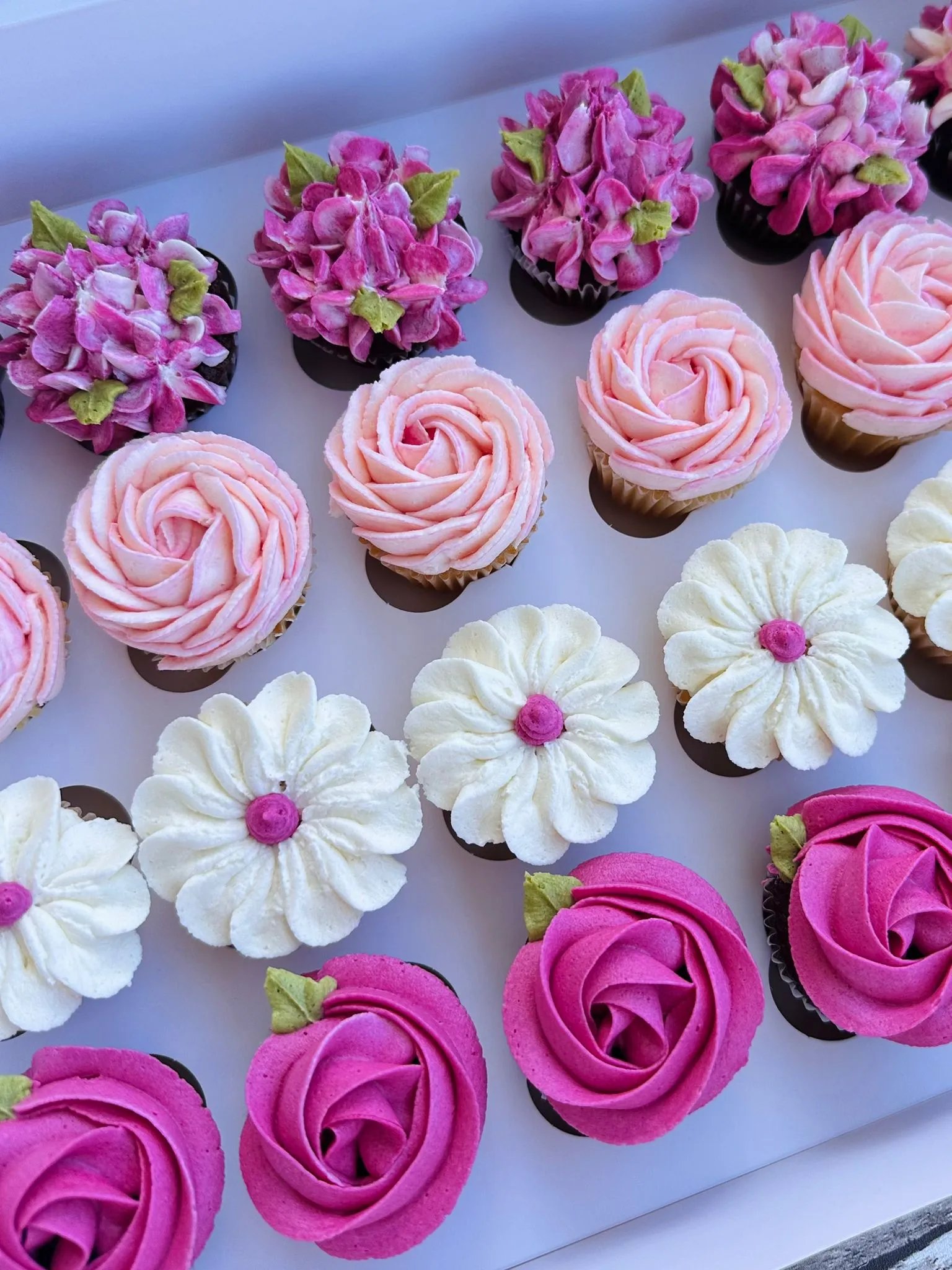 Box of assorted cupcakes decorated with pink, white, and purple flower-themed frosting.