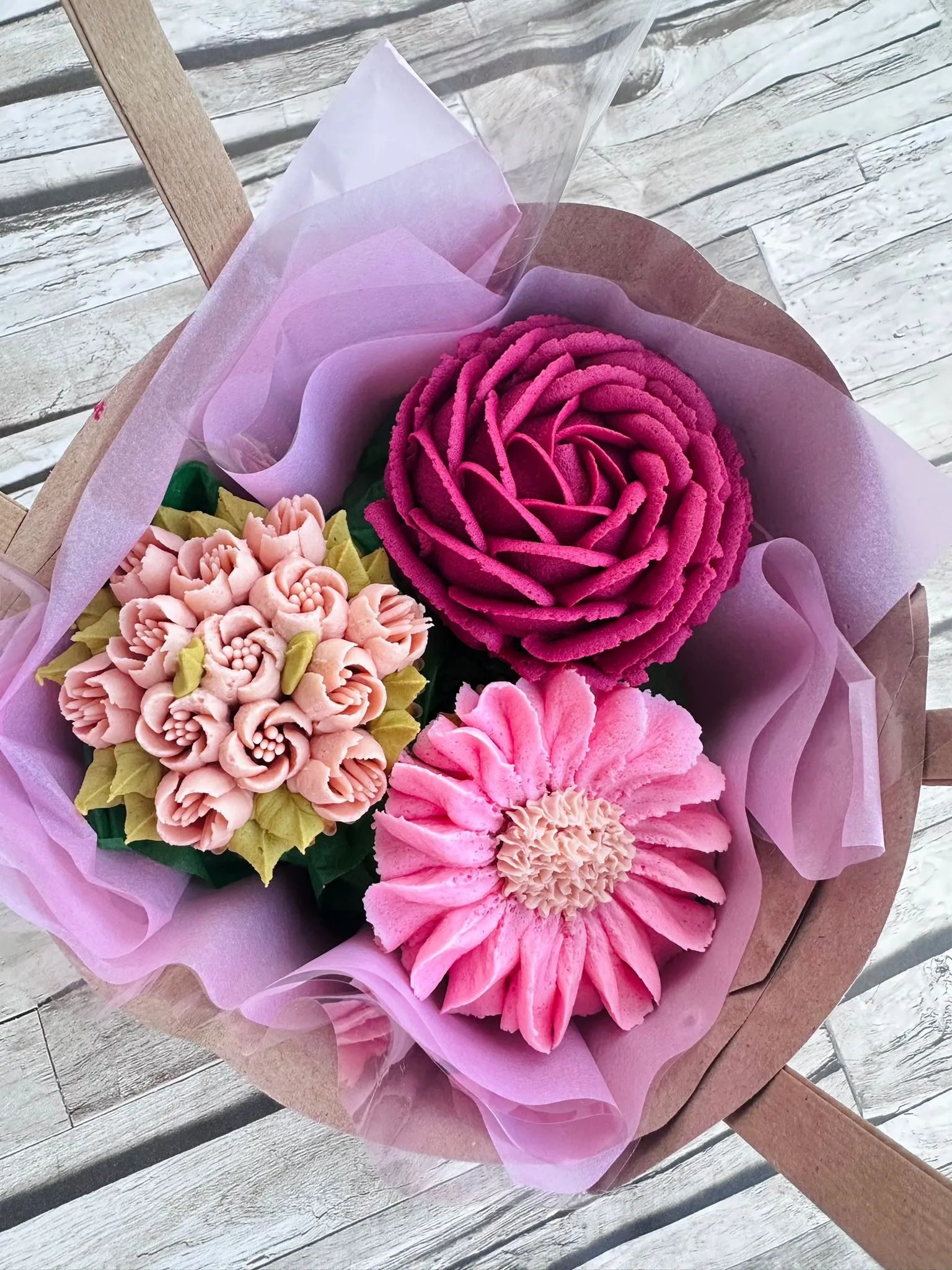 Bouquet of three detailed, colorful cake flowers in pink, fuchsia, and peach, wrapped in purple tissue paper on a wooden table.
