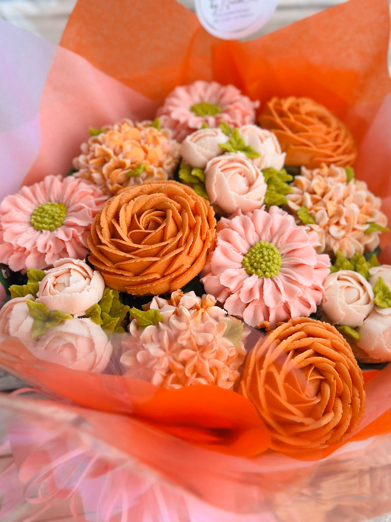 Bouquet of intricately piped buttercream flowers in shades of peach, pink, and orange with green leaves, wrapped in orange and pink tissue paper.