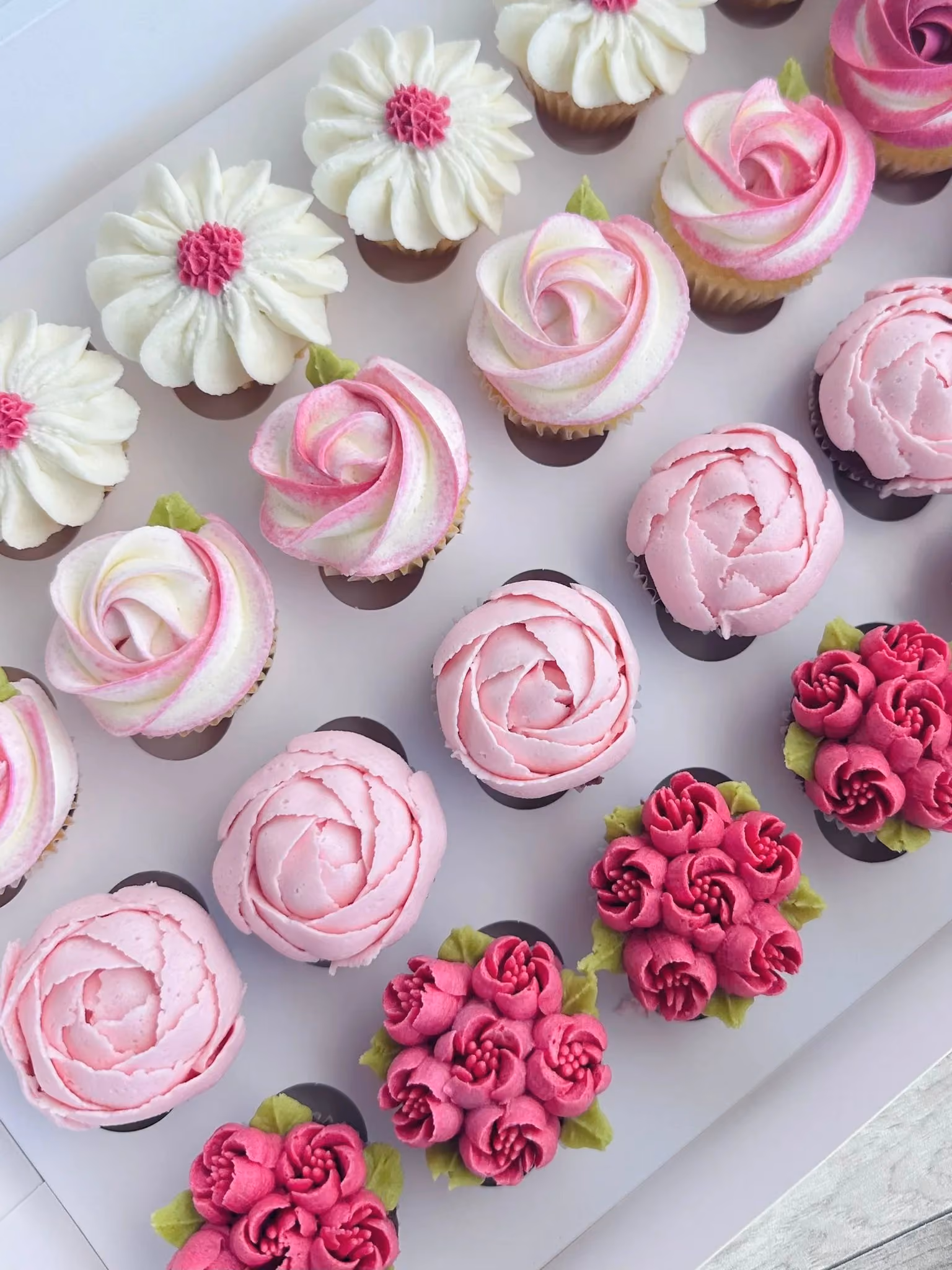 Assorted cupcakes decorated with buttercream flowers in shades of white, pink, and magenta arranged in a white box.
