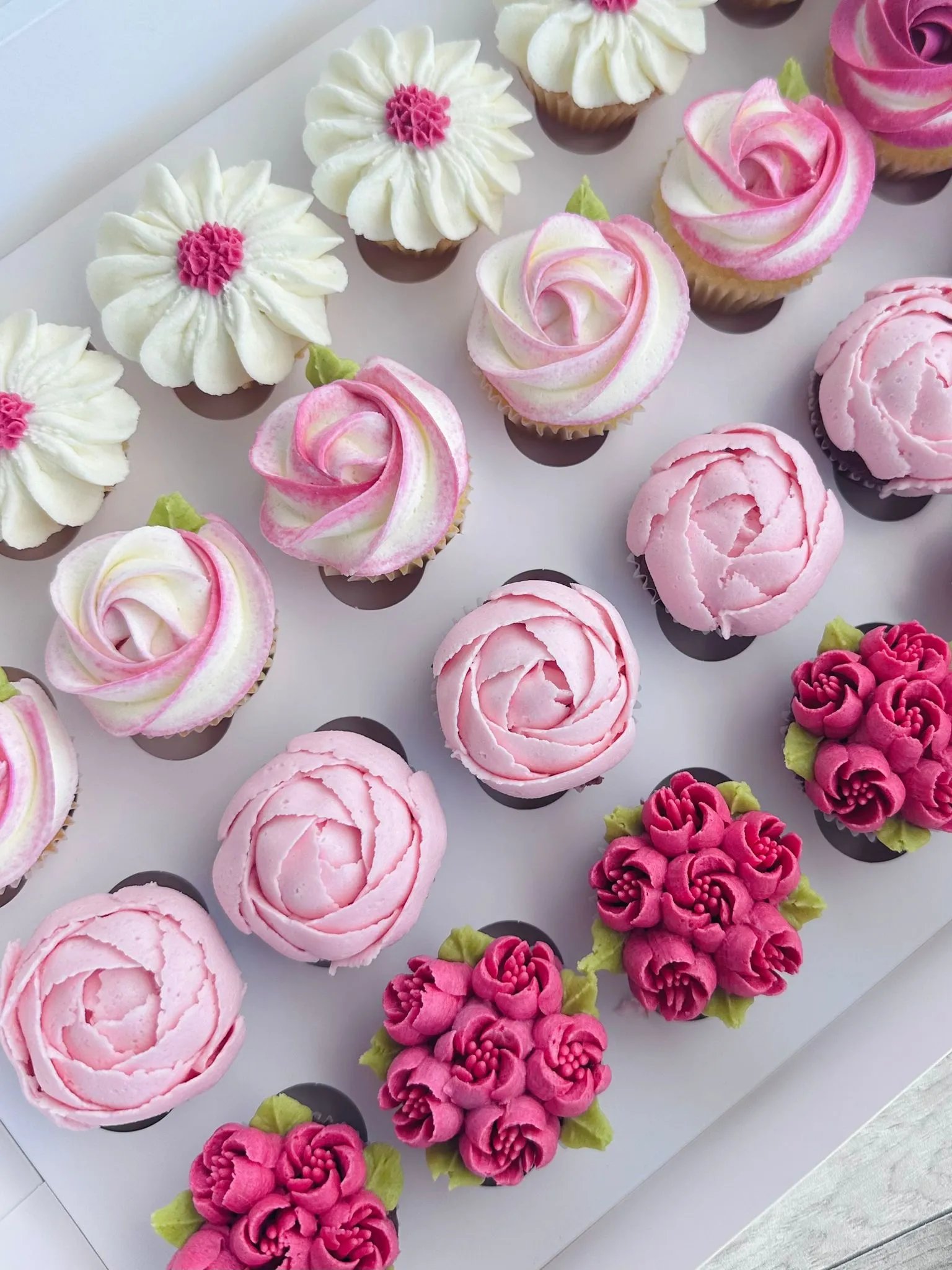 Assorted cupcakes decorated with buttercream flowers in shades of white, pink, and magenta arranged in a white box.
