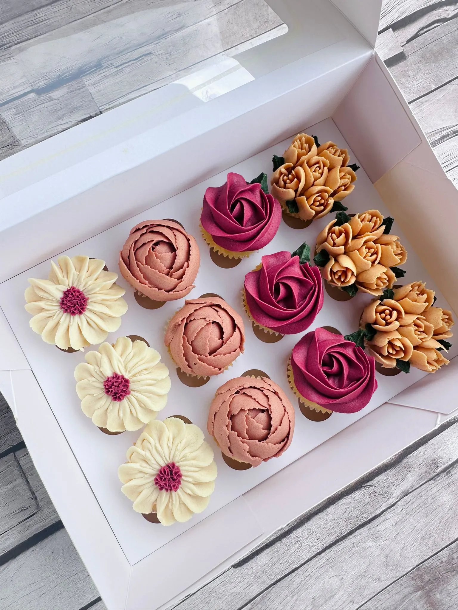 Box of a dozen assorted cupcakes decorated with buttercream flowers in white, pink, purple, and orange colors.
