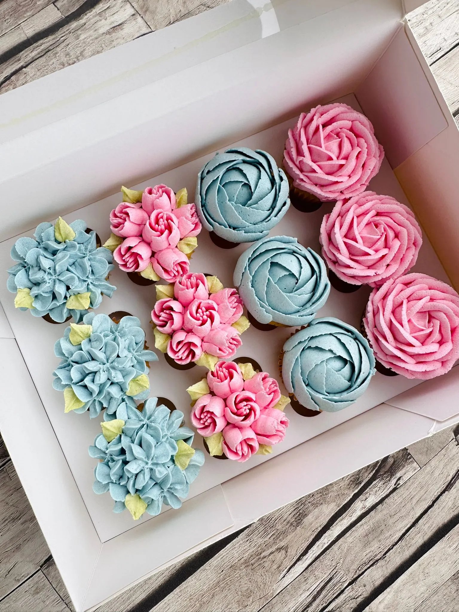Box of 12 cupcakes decorated with pink and blue floral buttercream designs, arranged in three rows on a wooden surface.