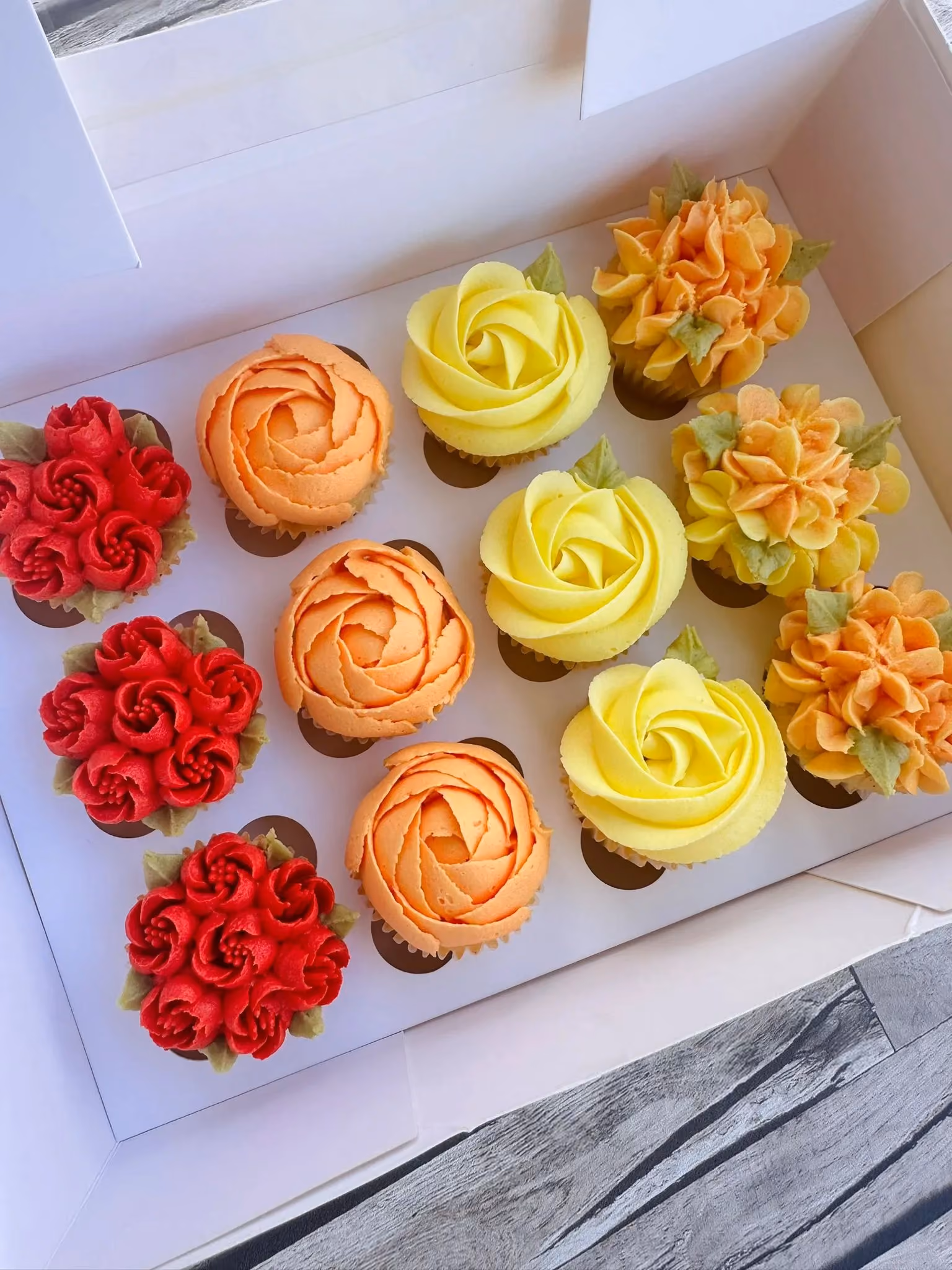 Box of twelve assorted cupcakes with pink, red, and white flower-shaped frosting decorations accented with green leaves.