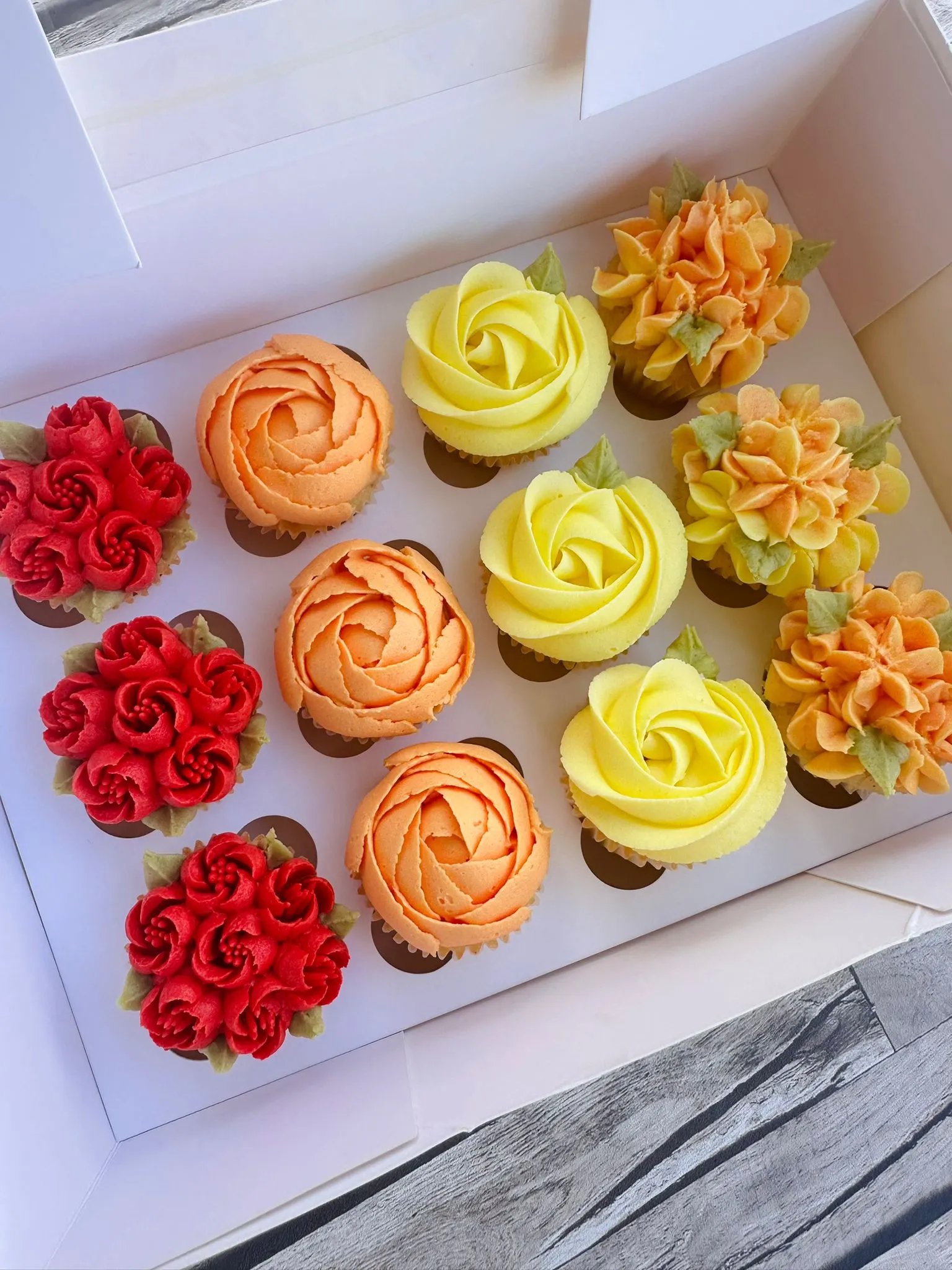 Box of twelve assorted cupcakes with pink, red, and white flower-shaped frosting decorations accented with green leaves.