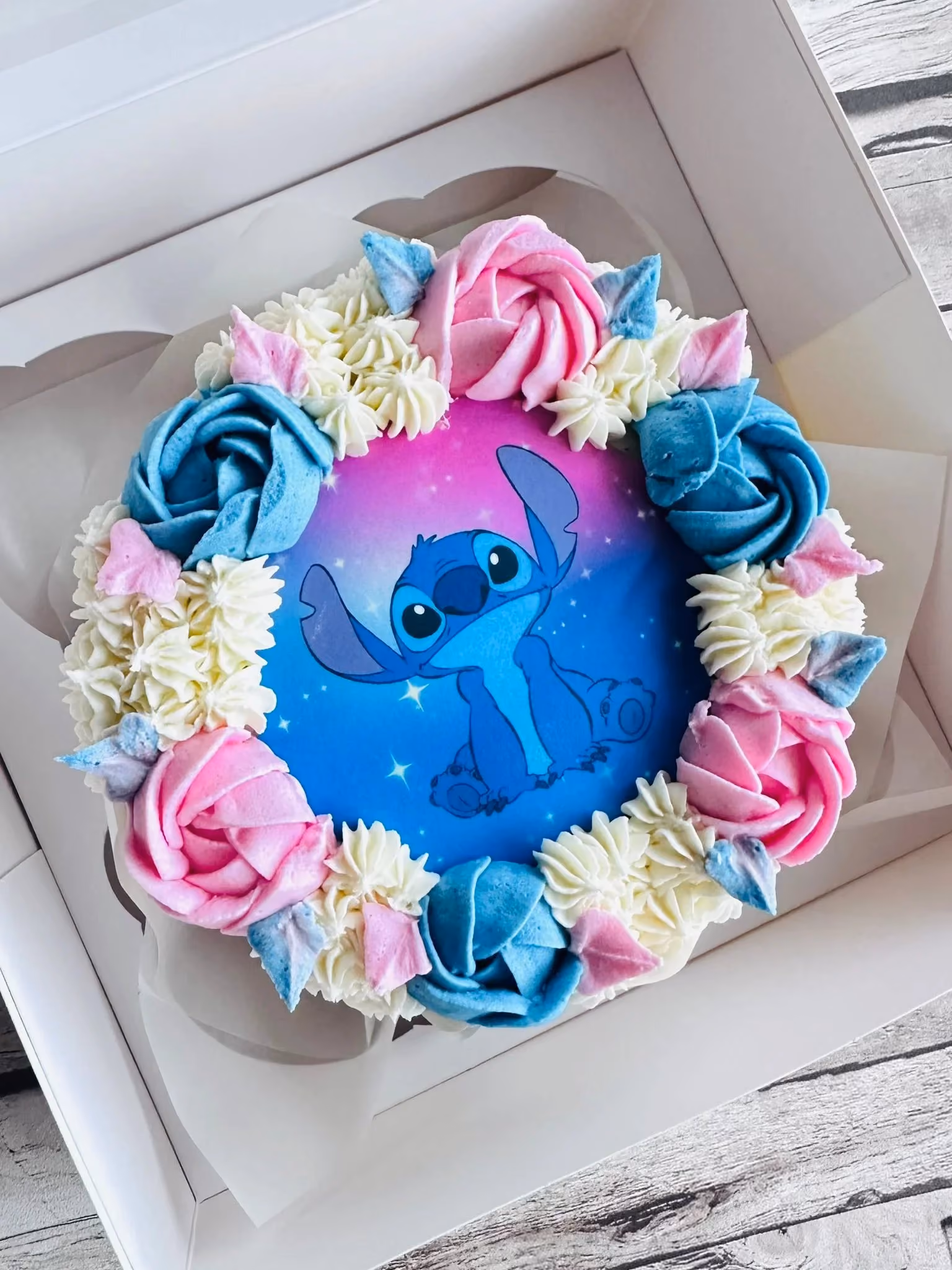 Round cake with blue and pink frosting roses and white cream, featuring a printed image of Stitch from Lilo & Stitch on a purple and blue gradient background.