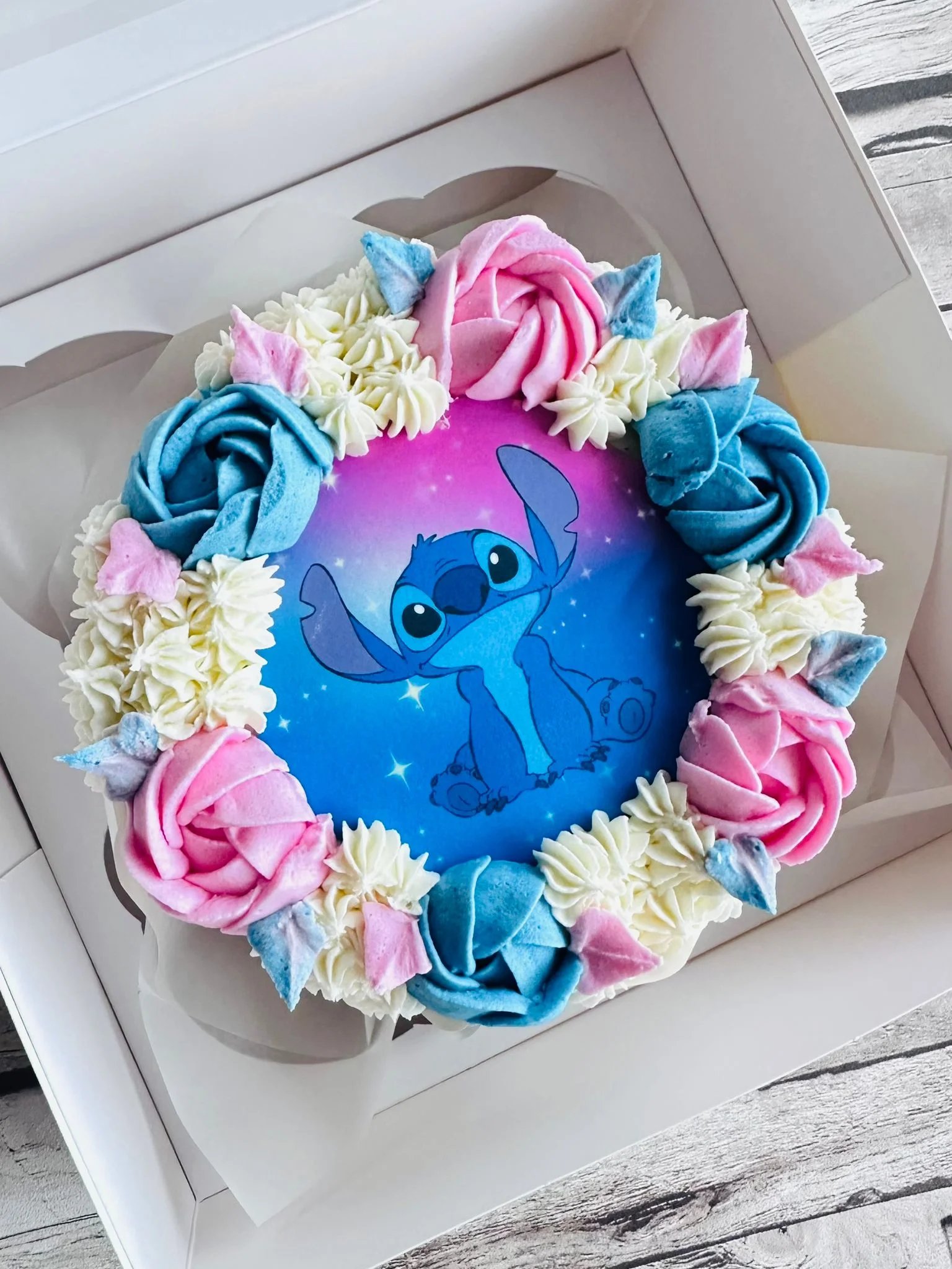 Round cake with blue and pink frosting roses and white cream, featuring a printed image of Stitch from Lilo & Stitch on a purple and blue gradient background.