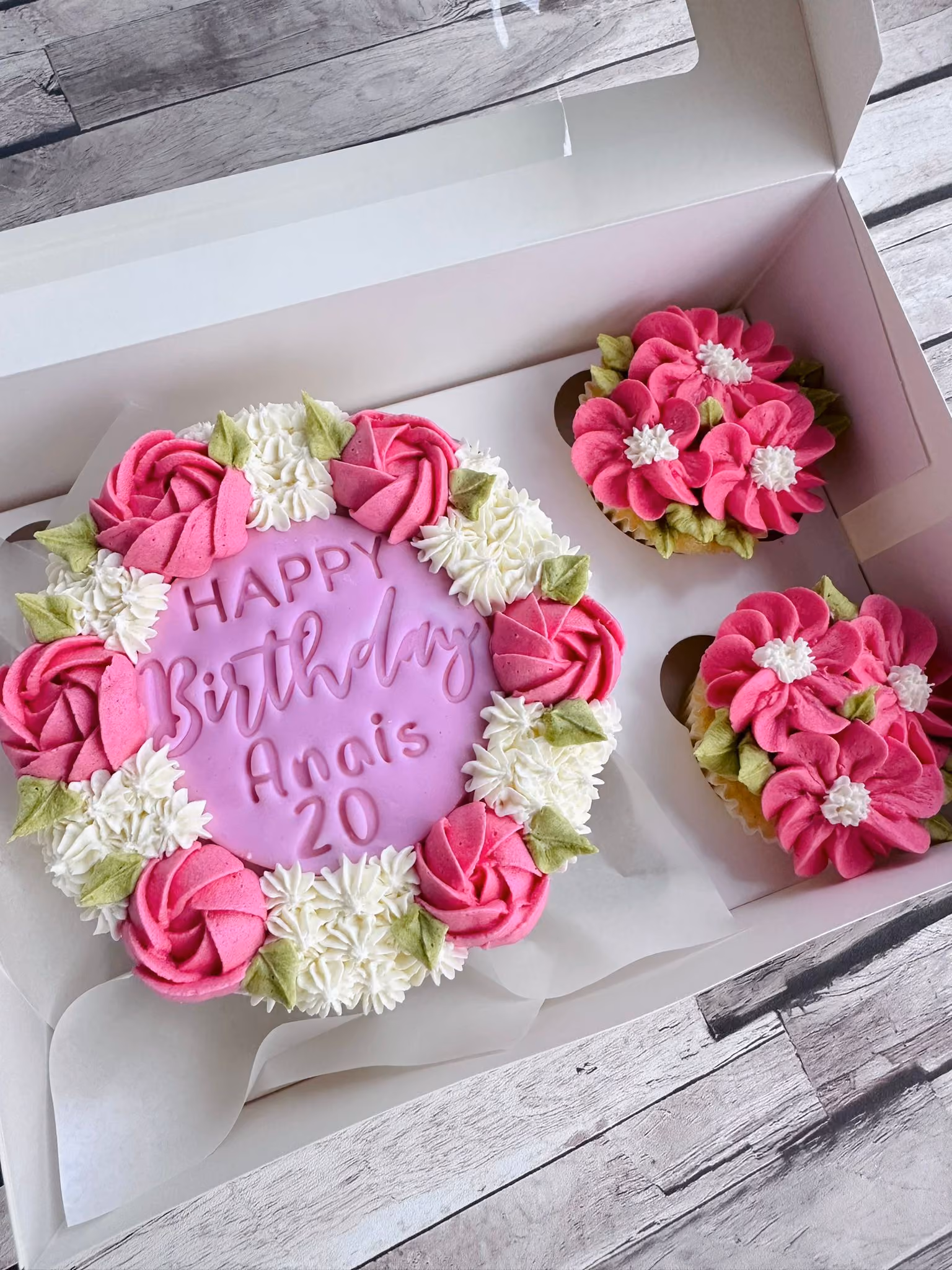 Round birthday cake with pink and white floral icing and the message 'Happy Birthday Anais 20', with two matching floral cupcakes in a white box.