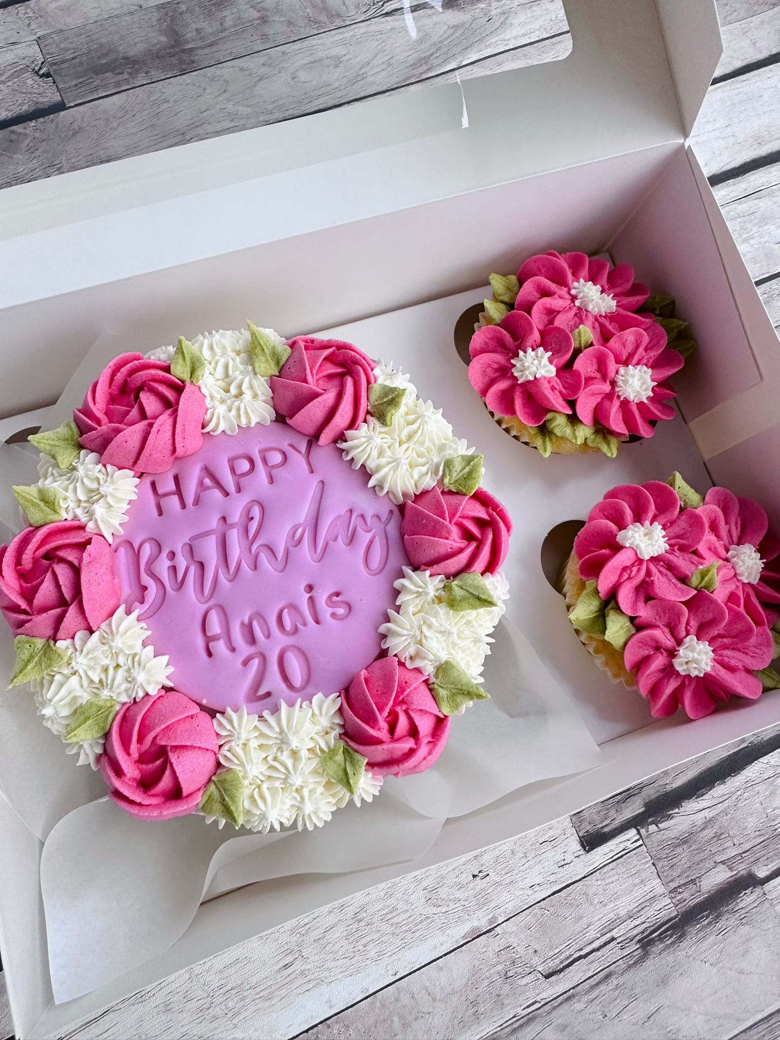 Round birthday cake with pink and white floral icing and the message 'Happy Birthday Anais 20', with two matching floral cupcakes in a white box.