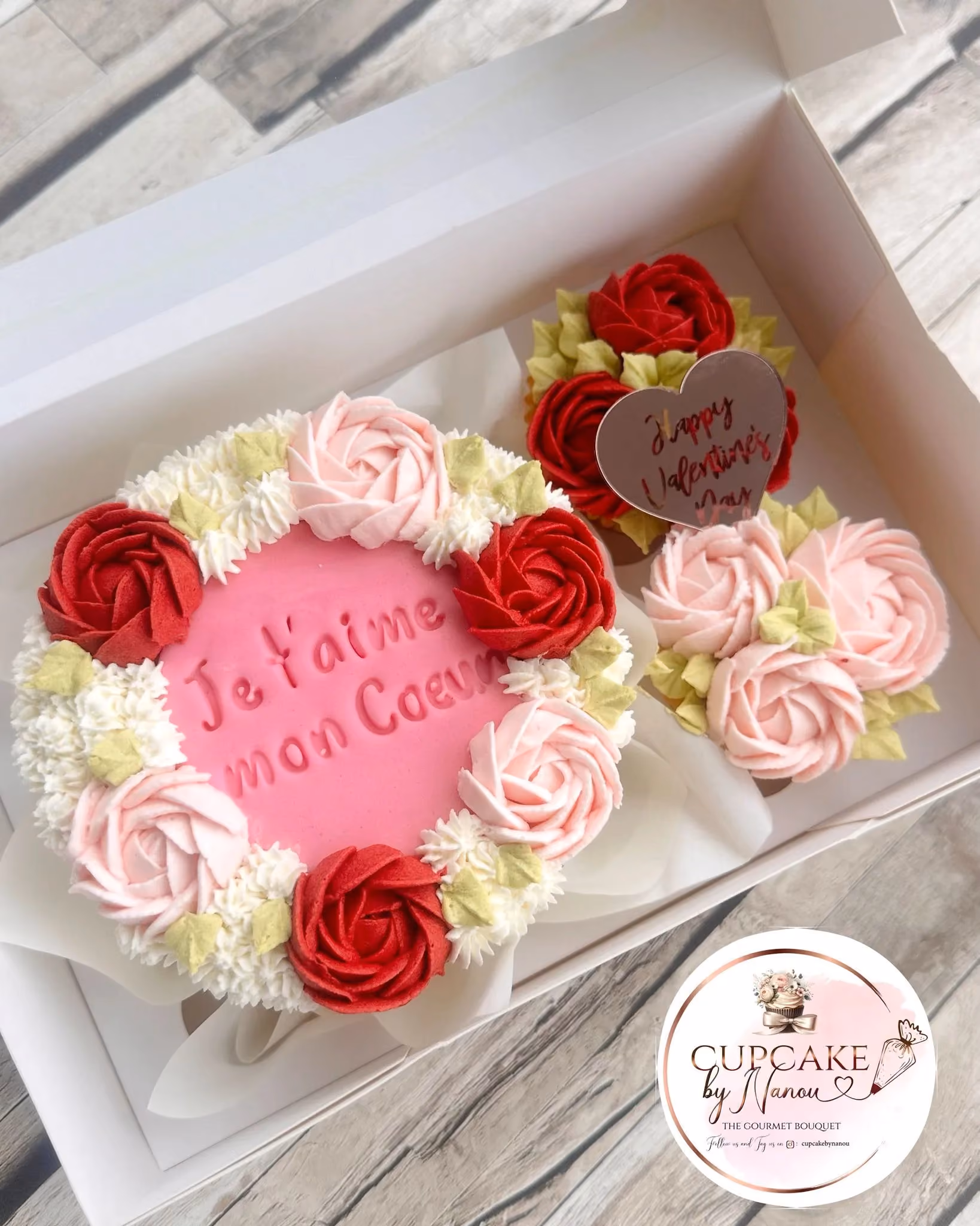 Heart-shaped pink cake decorated with red, pink, and white buttercream roses and green leaves, with the French text 'Je t'aime mon Coeur' on top, accompanied by two smaller cupcakes with rose decorations and a heart-shaped tag reading 'Happy Valentine's Day'.