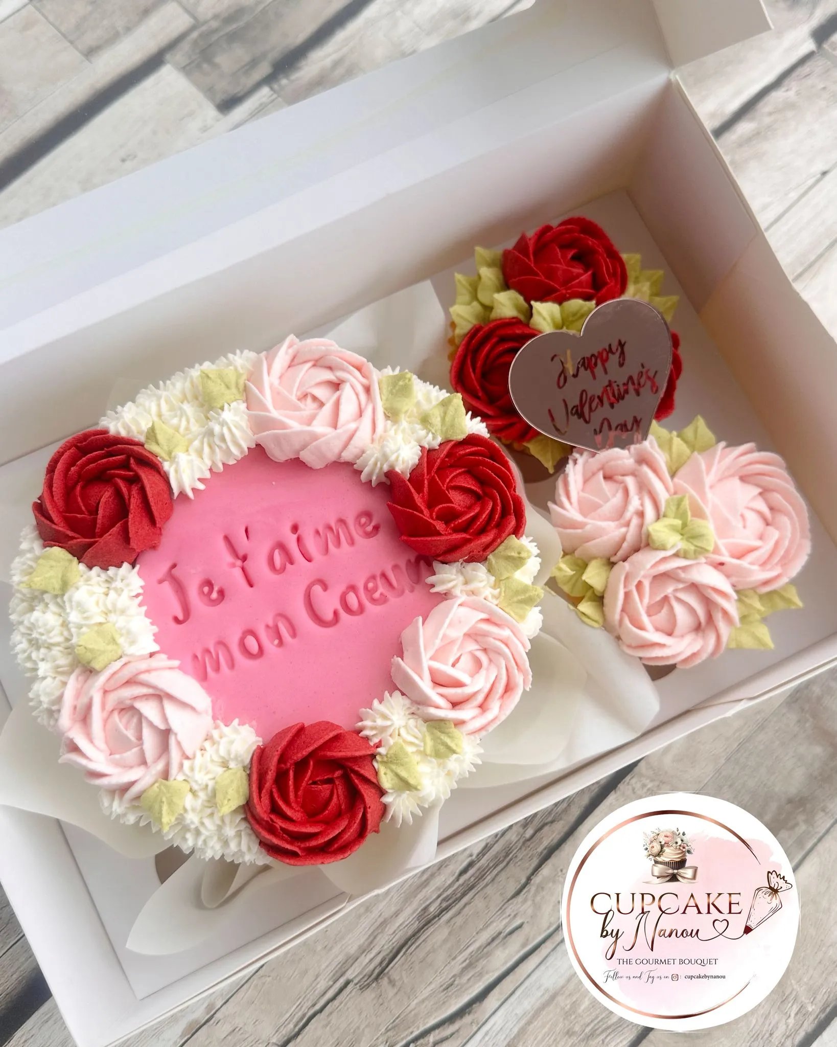 Heart-shaped pink cake decorated with red, pink, and white buttercream roses and green leaves, with the French text 'Je t'aime mon Coeur' on top, accompanied by two smaller cupcakes with rose decorations and a heart-shaped tag reading 'Happy Valentine's Day'.