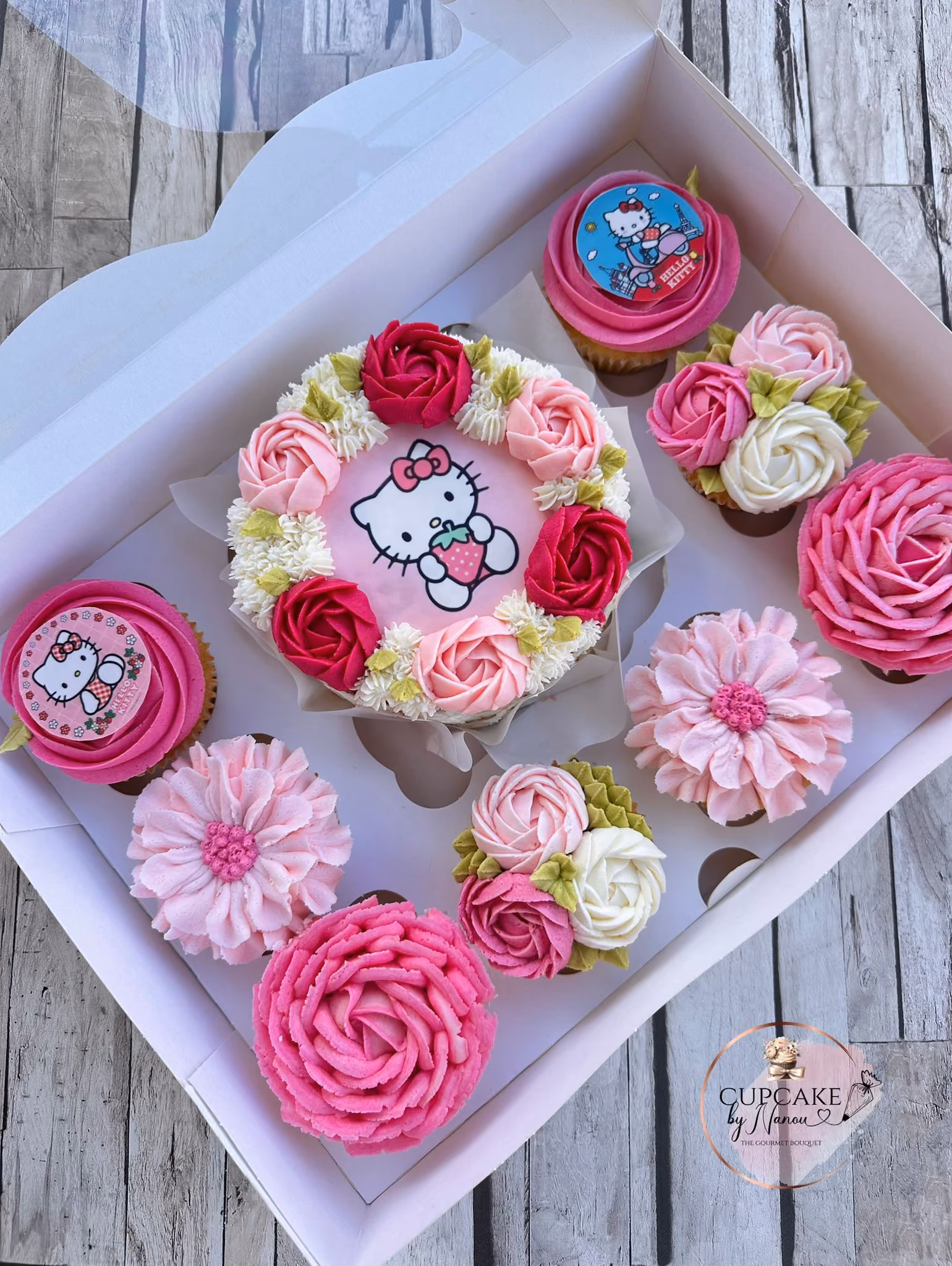 Box of cupcakes and a small cake decorated with pink, white, and red floral frosting and Hello Kitty edible images.