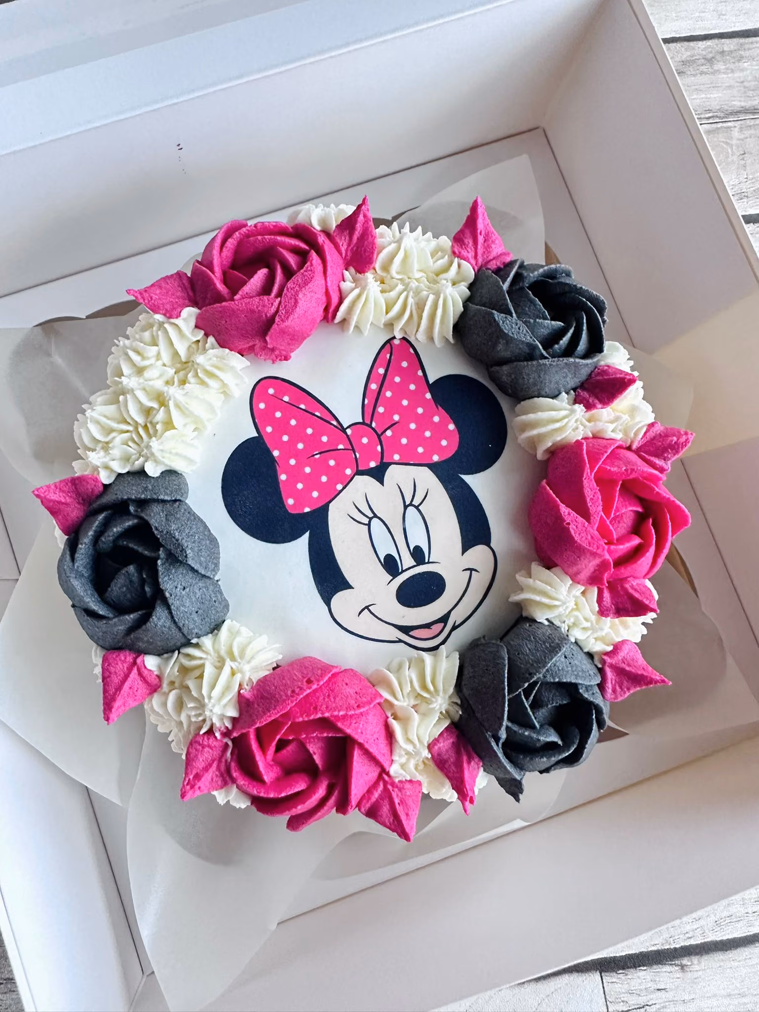 Round cake decorated with pink and gray icing roses and white swirls, featuring a Minnie Mouse face with a pink polka dot bow in the center.