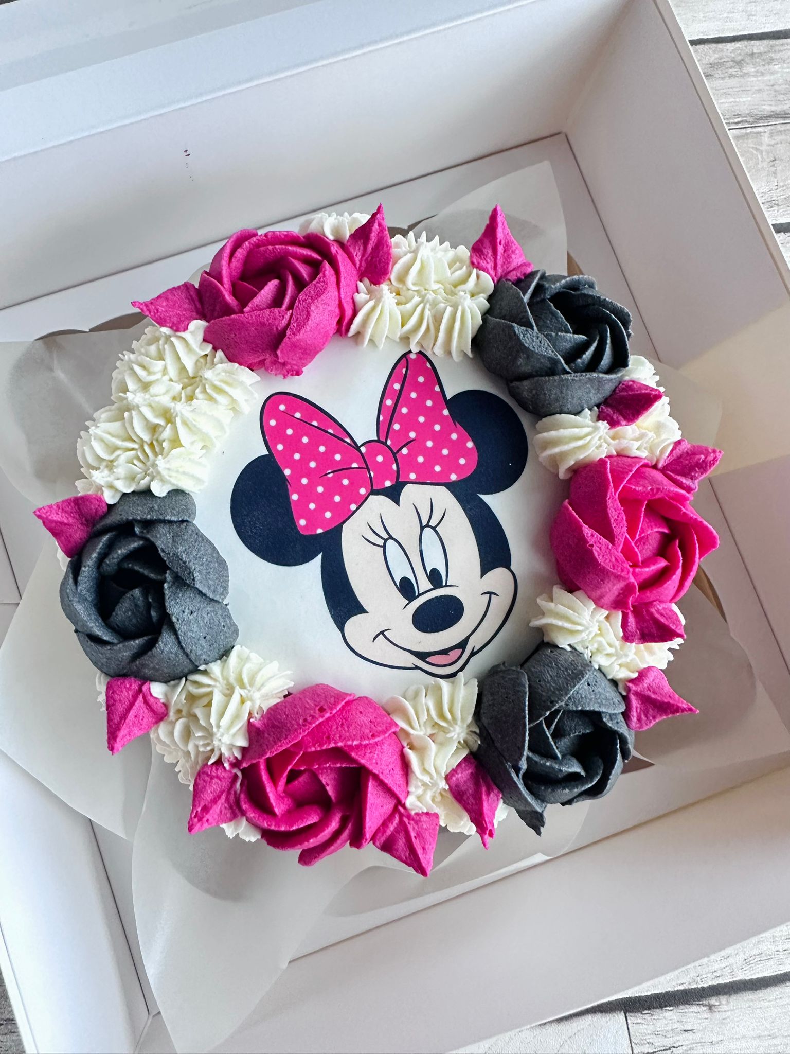 Round cake decorated with pink and gray icing roses and white swirls, featuring a Minnie Mouse face with a pink polka dot bow in the center.