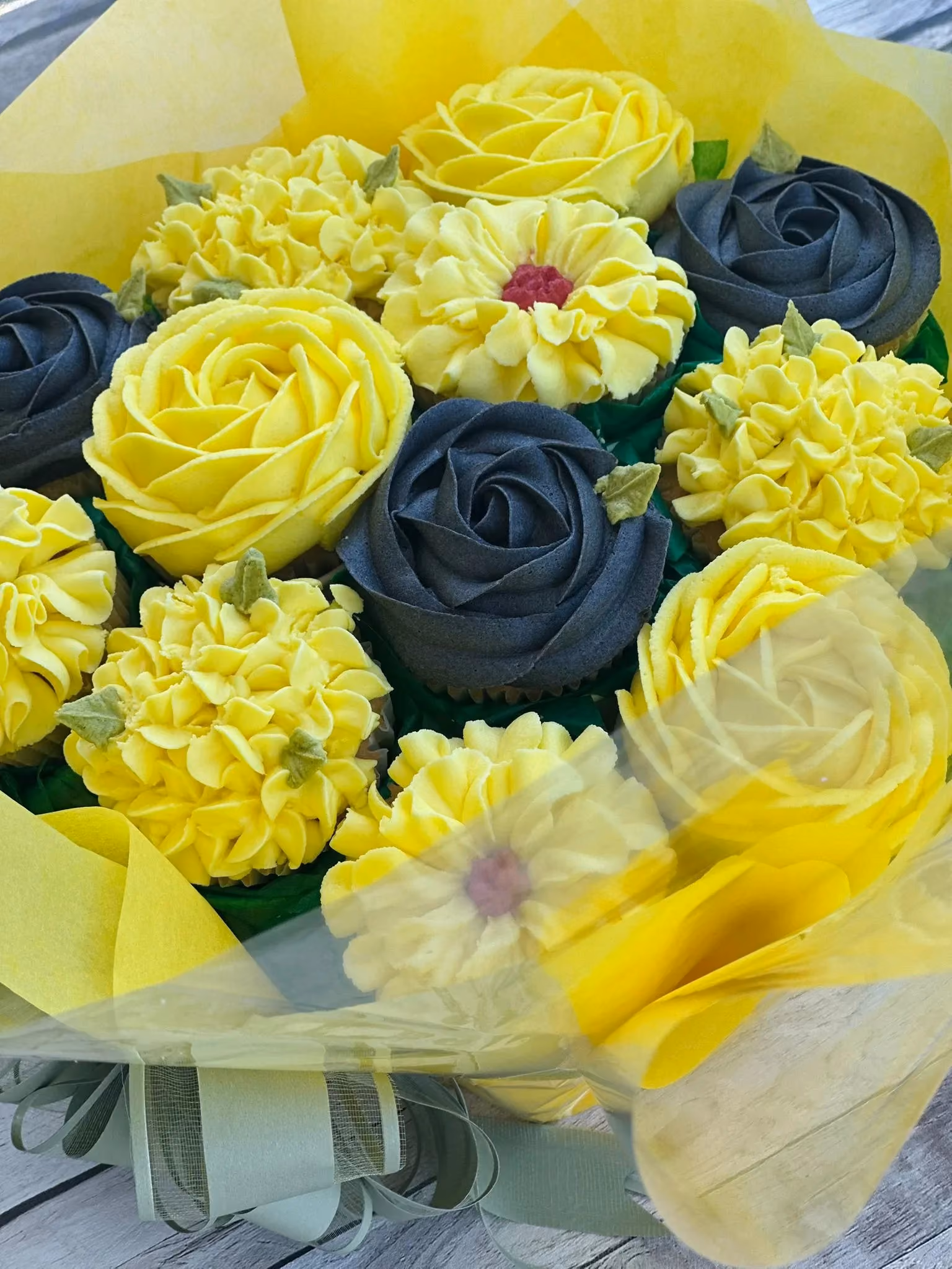 Bouquet of cupcakes decorated with yellow and dark gray frosting designed to resemble flowers, wrapped in yellow paper with a green ribbon.