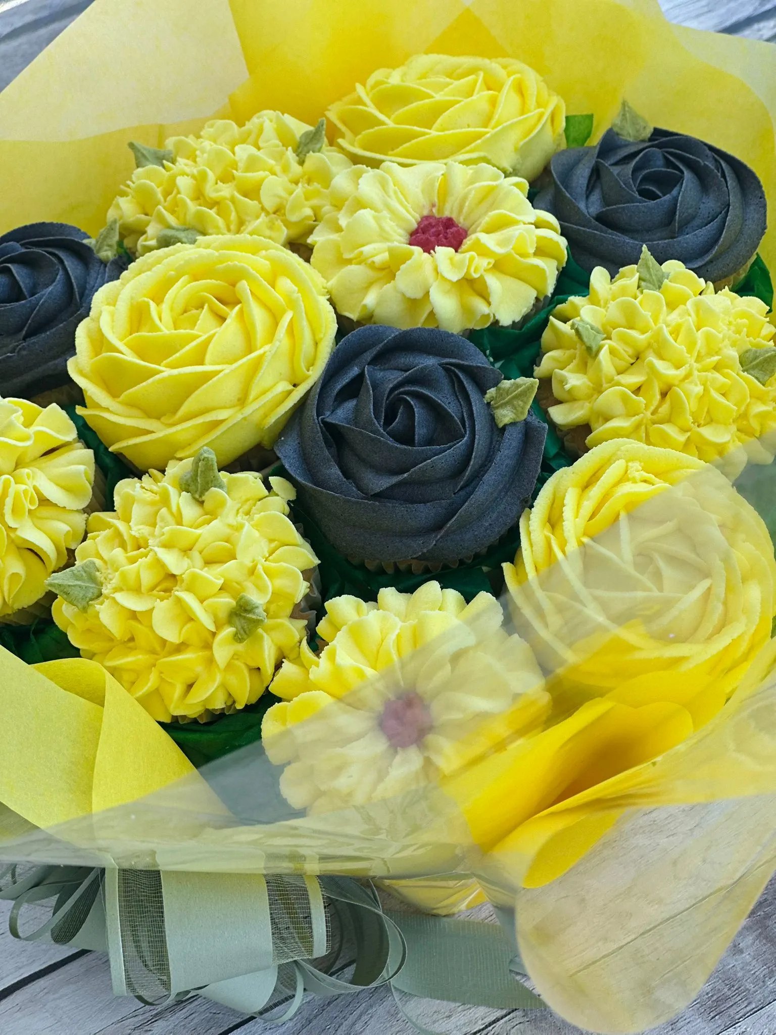 Bouquet of cupcakes decorated with yellow and dark gray frosting designed to resemble flowers, wrapped in yellow paper with a green ribbon.