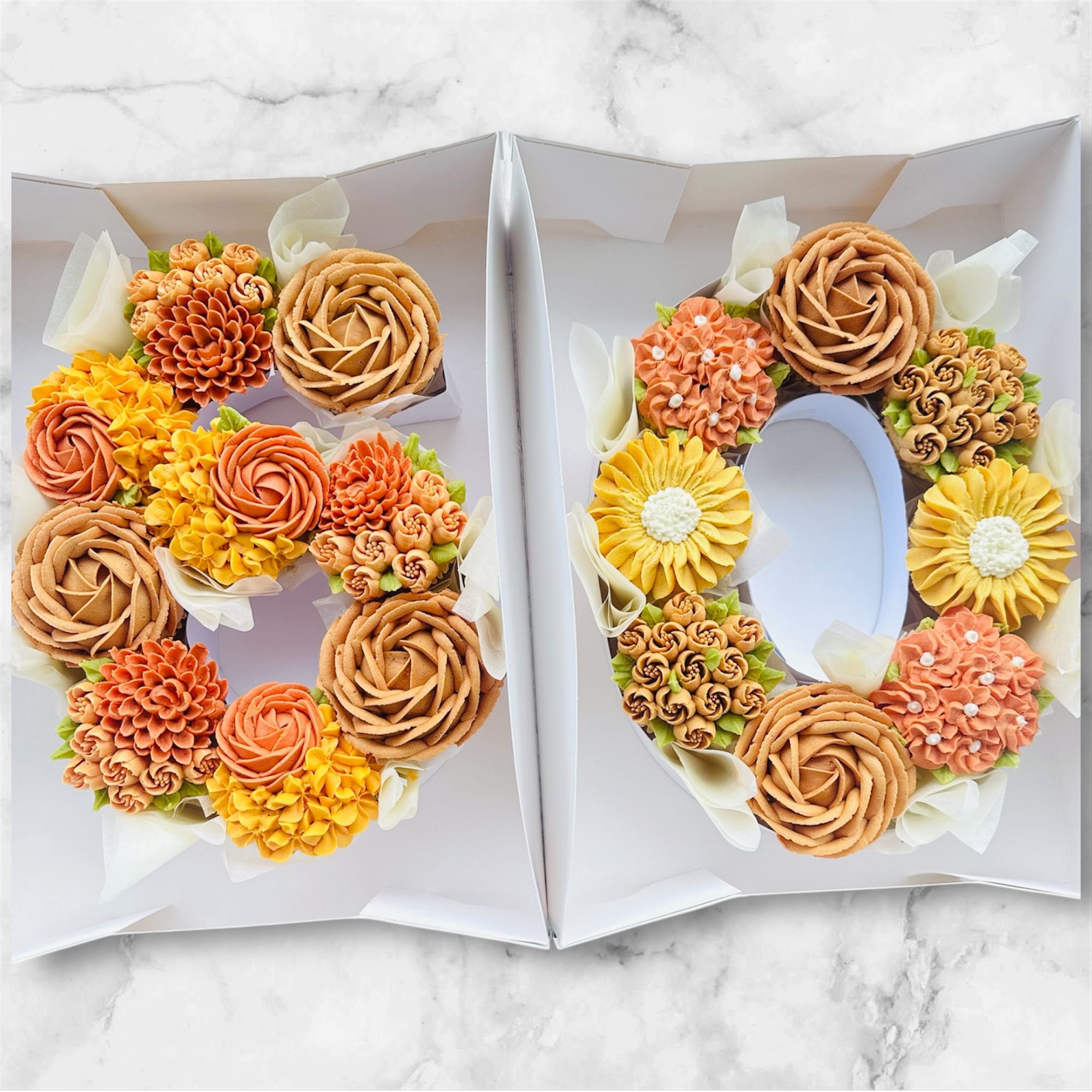 Two white boxes containing cupcakes arranged to form the numbers 5 and 0 decorated with orange, yellow, and beige flower-shaped frosting.