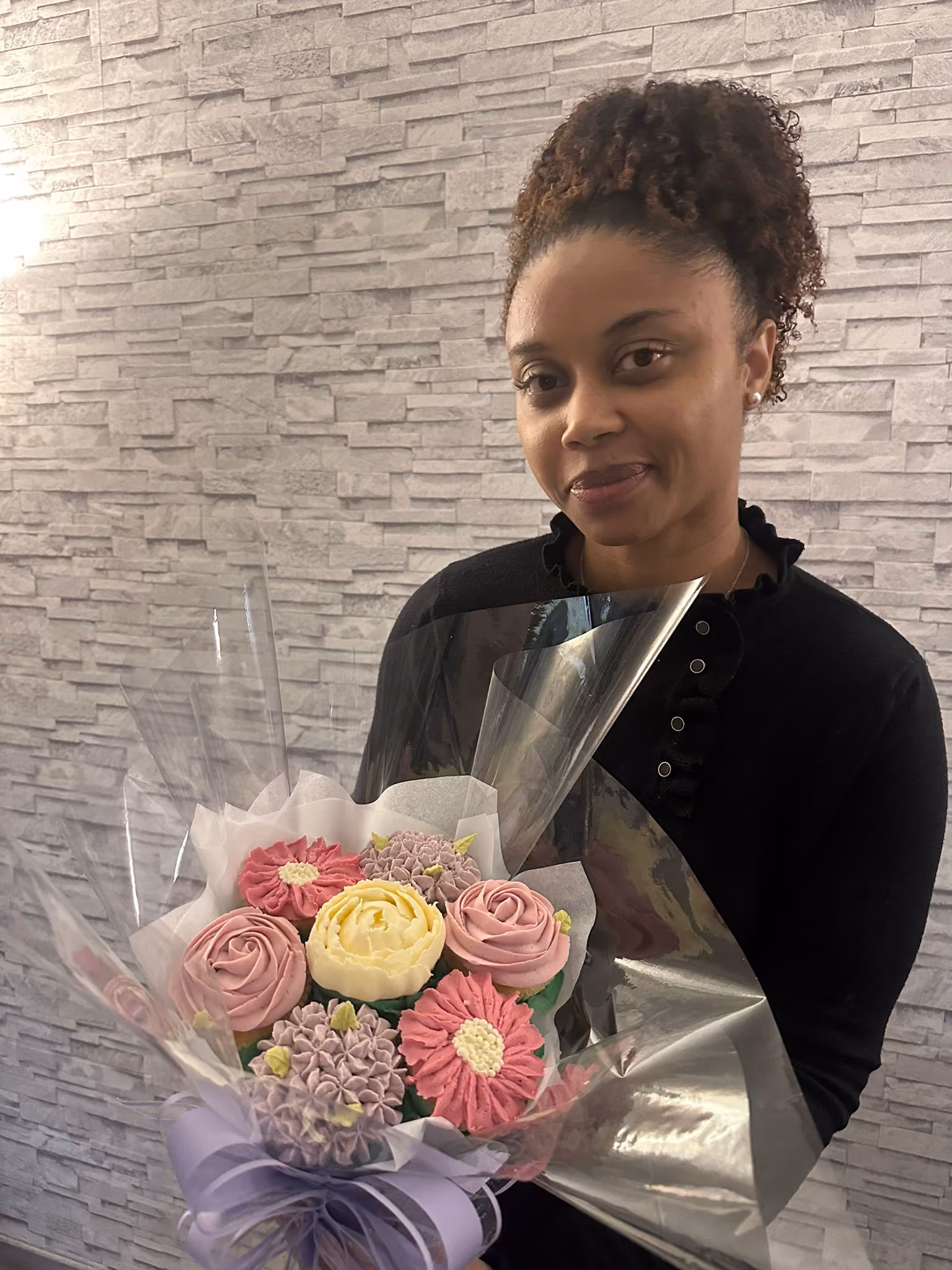 Woman wearing a black top holding a bouquet of pastel-colored flower cupcakes wrapped in clear plastic with a lavender ribbon.