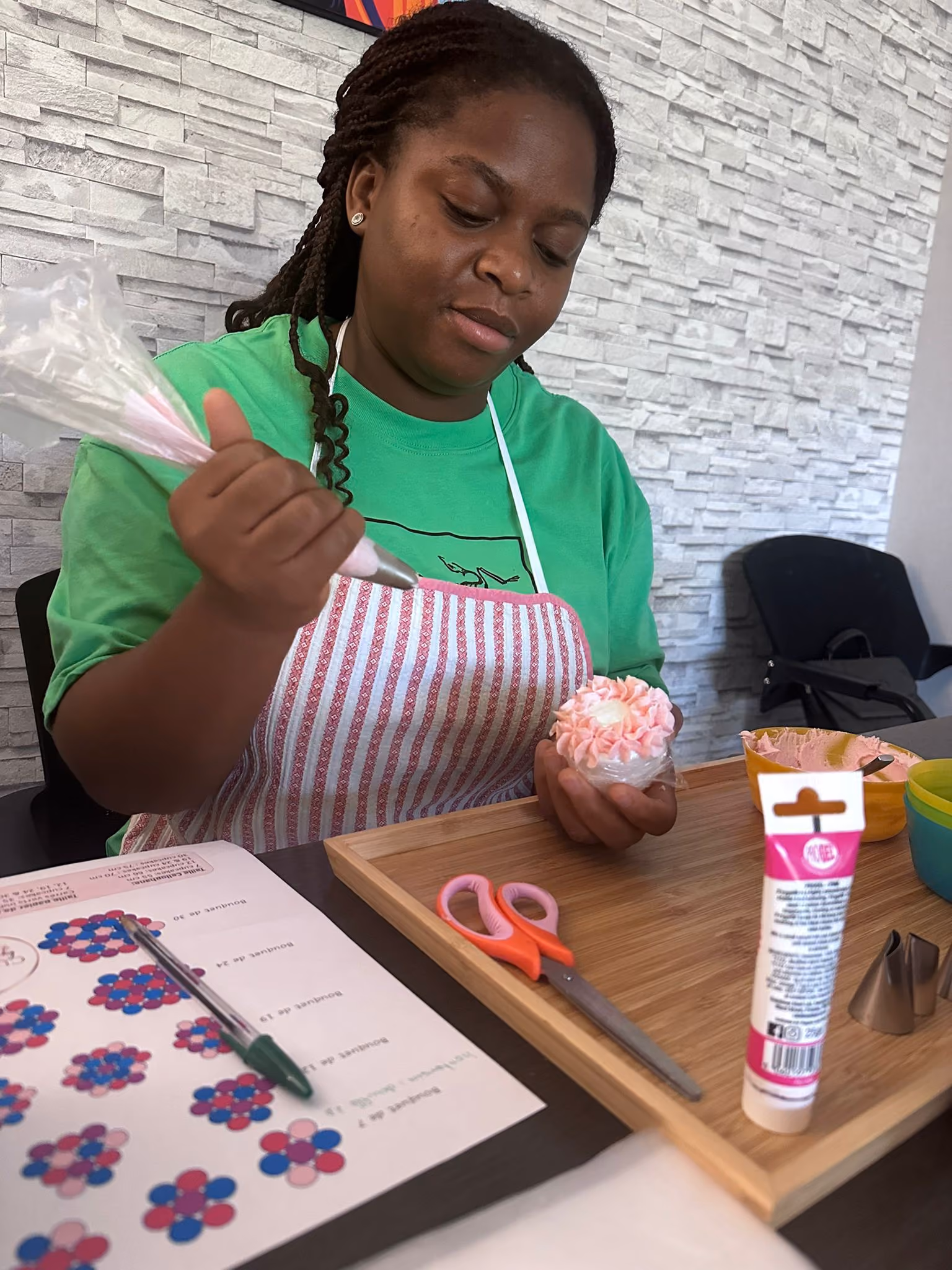 Woman in a green shirt and striped apron decorating a cupcake with pink frosting using a piping bag at a table with decorating tools and instructions.
