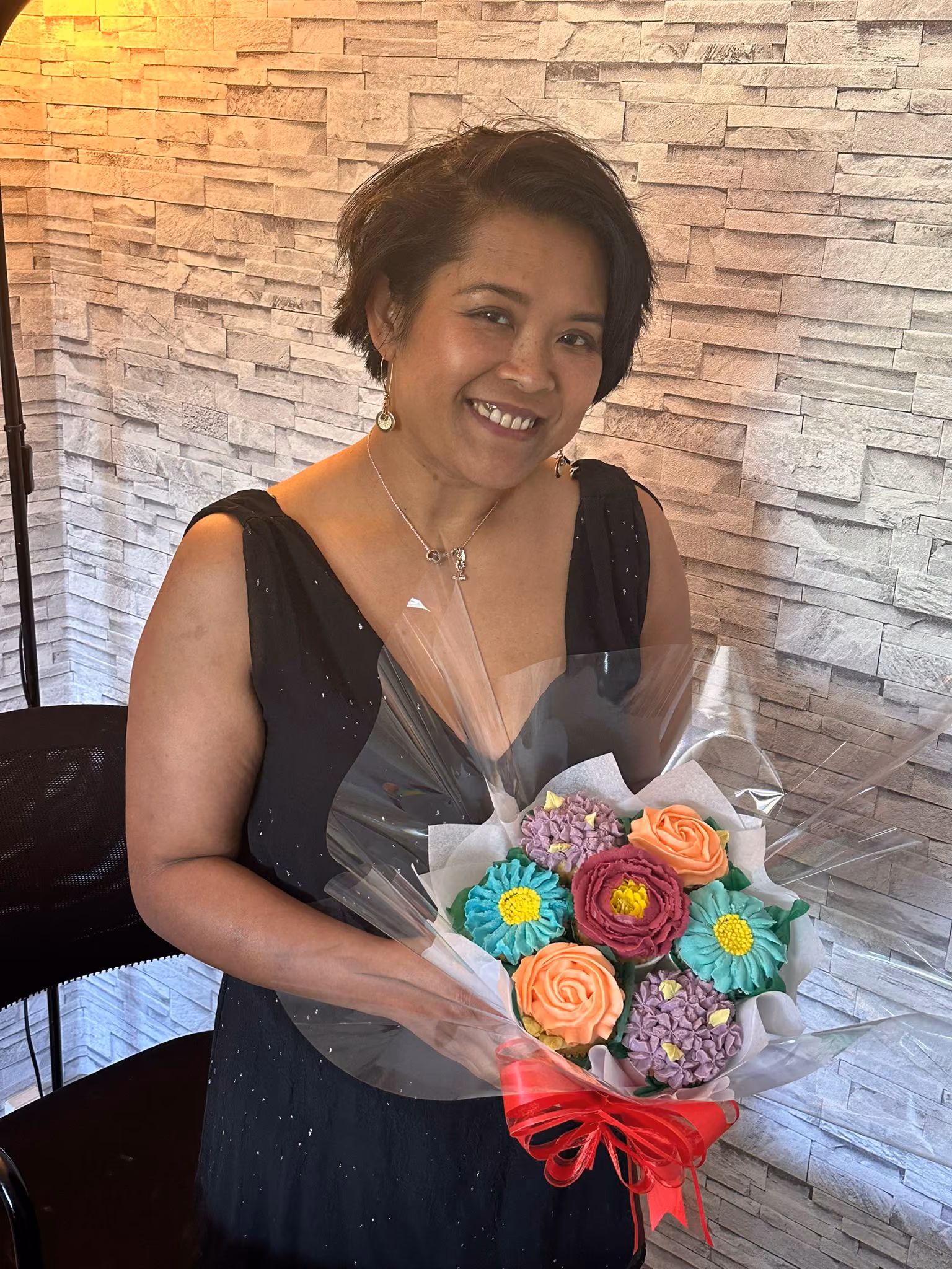 Smiling woman in a black sleeveless dress holding a bouquet of colorful flower-shaped cupcakes wrapped in clear plastic with a red ribbon.