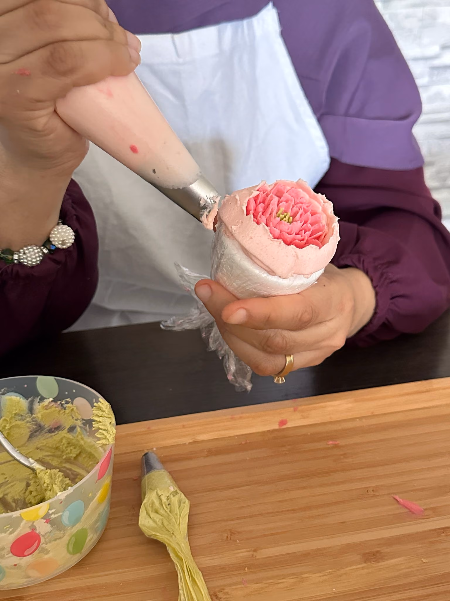 Person decorating a pink buttercream flower with a piping bag, wearing a white apron and purple sleeves.