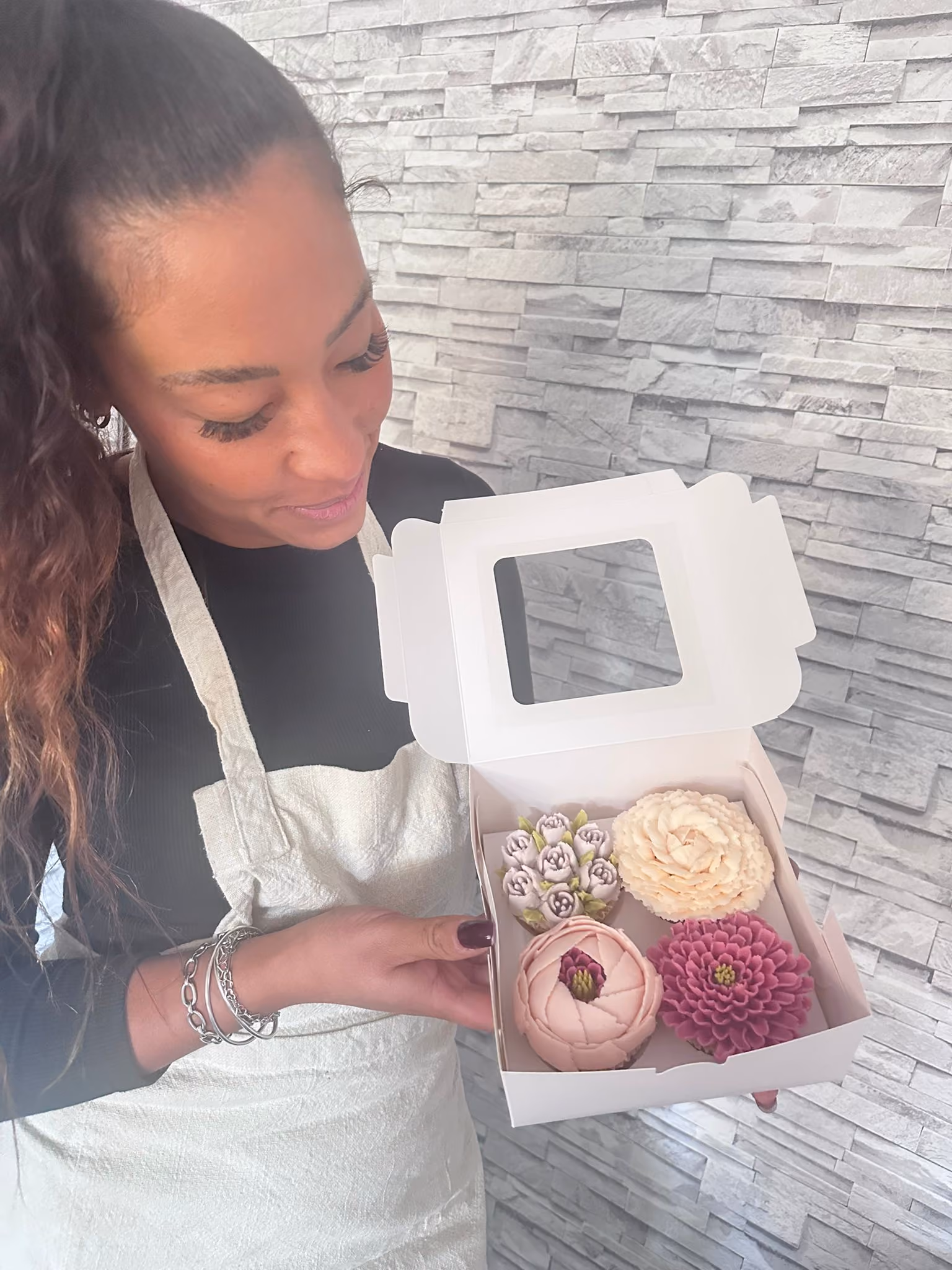 Woman wearing an apron holding a box containing four intricately decorated flower cupcakes against a textured stone wall.