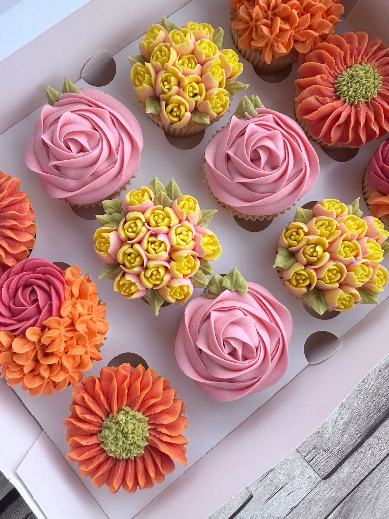 Box of assorted decorated cupcakes with pink rose, yellow flower clusters, and orange floral designs on a wooden surface.