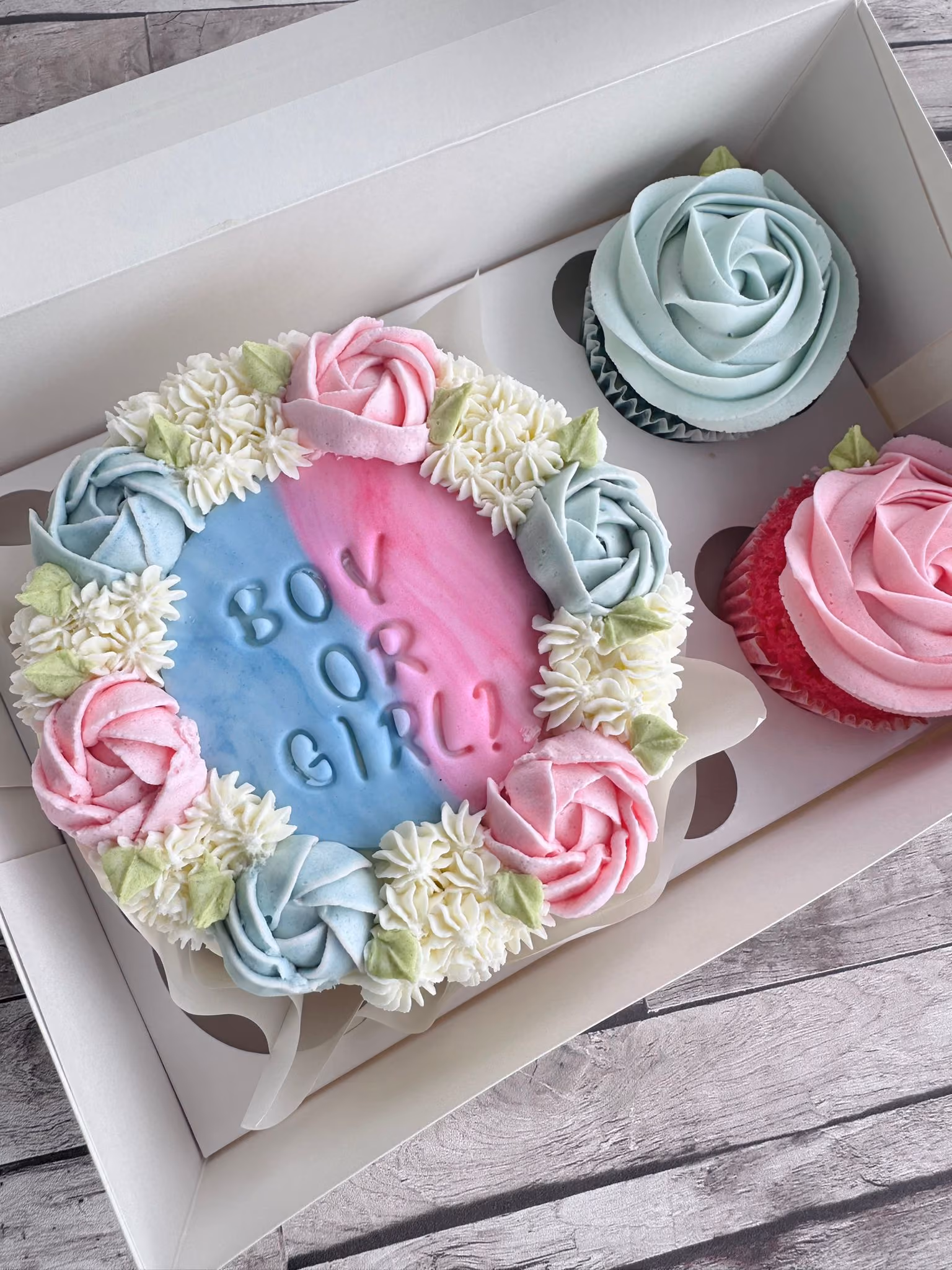 Gender reveal cake and two matching cupcakes with pink and blue frosting, cake decorated with flowers and text saying 'Boy or Girl?'.