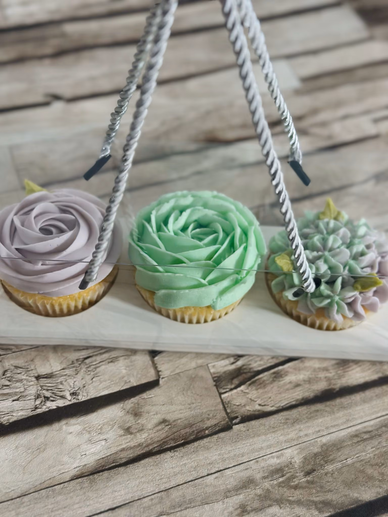 Three decorated cupcakes with pastel-colored floral frosting in a wooden holder on a wooden surface.