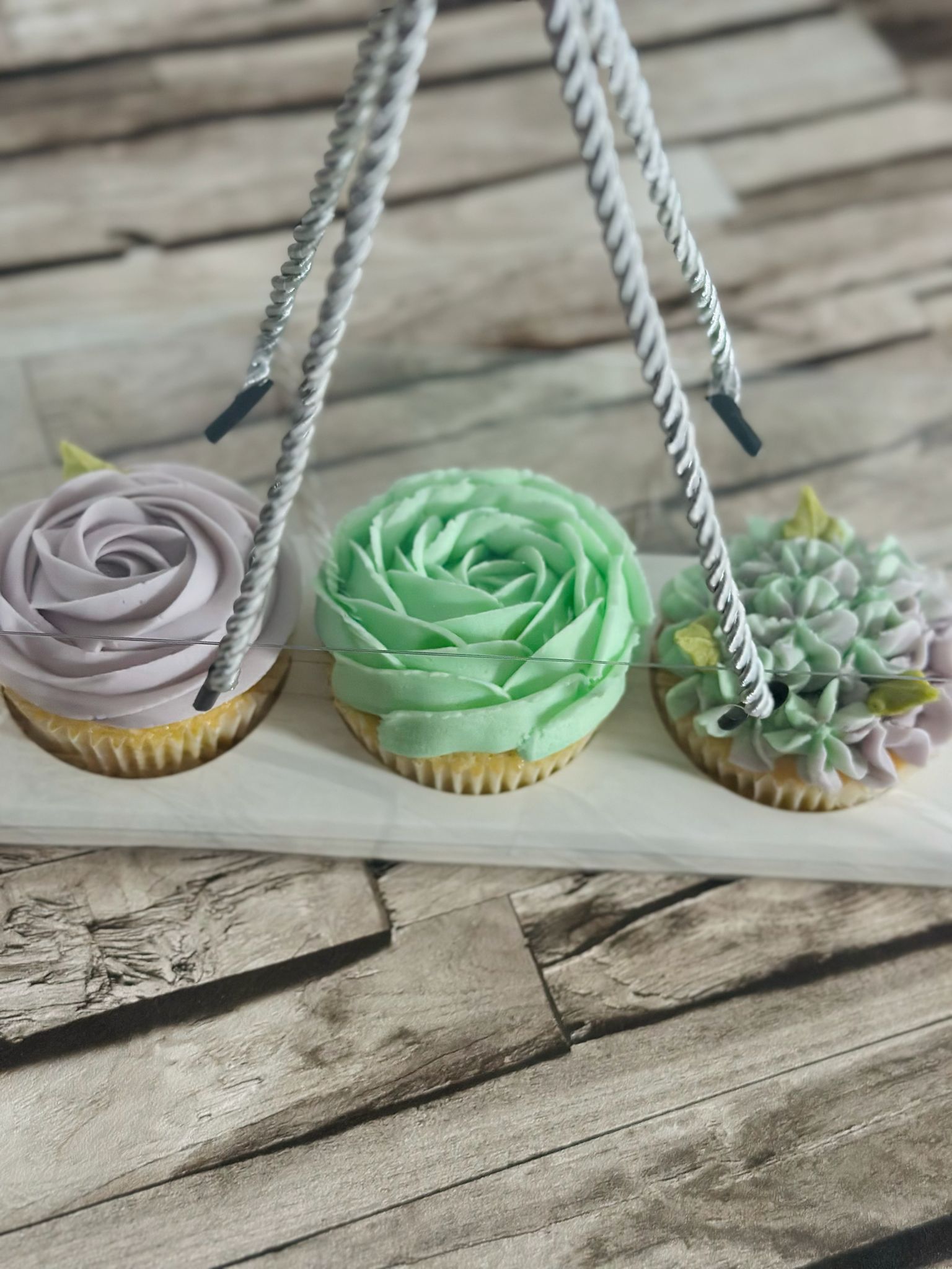 Three decorated cupcakes with pastel-colored floral frosting in a wooden holder on a wooden surface.