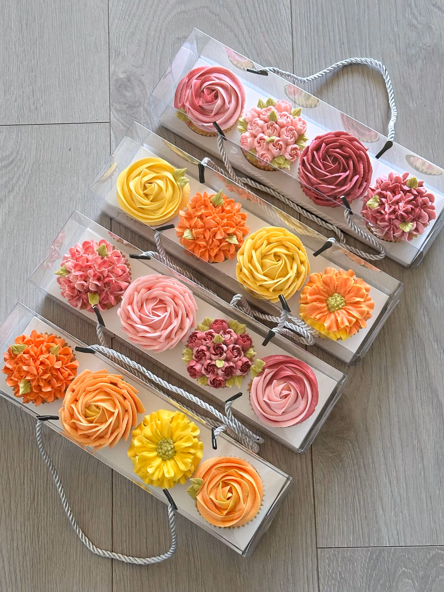 Four clear boxes each containing four cupcakes decorated with colorful floral frosting designs in pink, orange, yellow, and red shades.