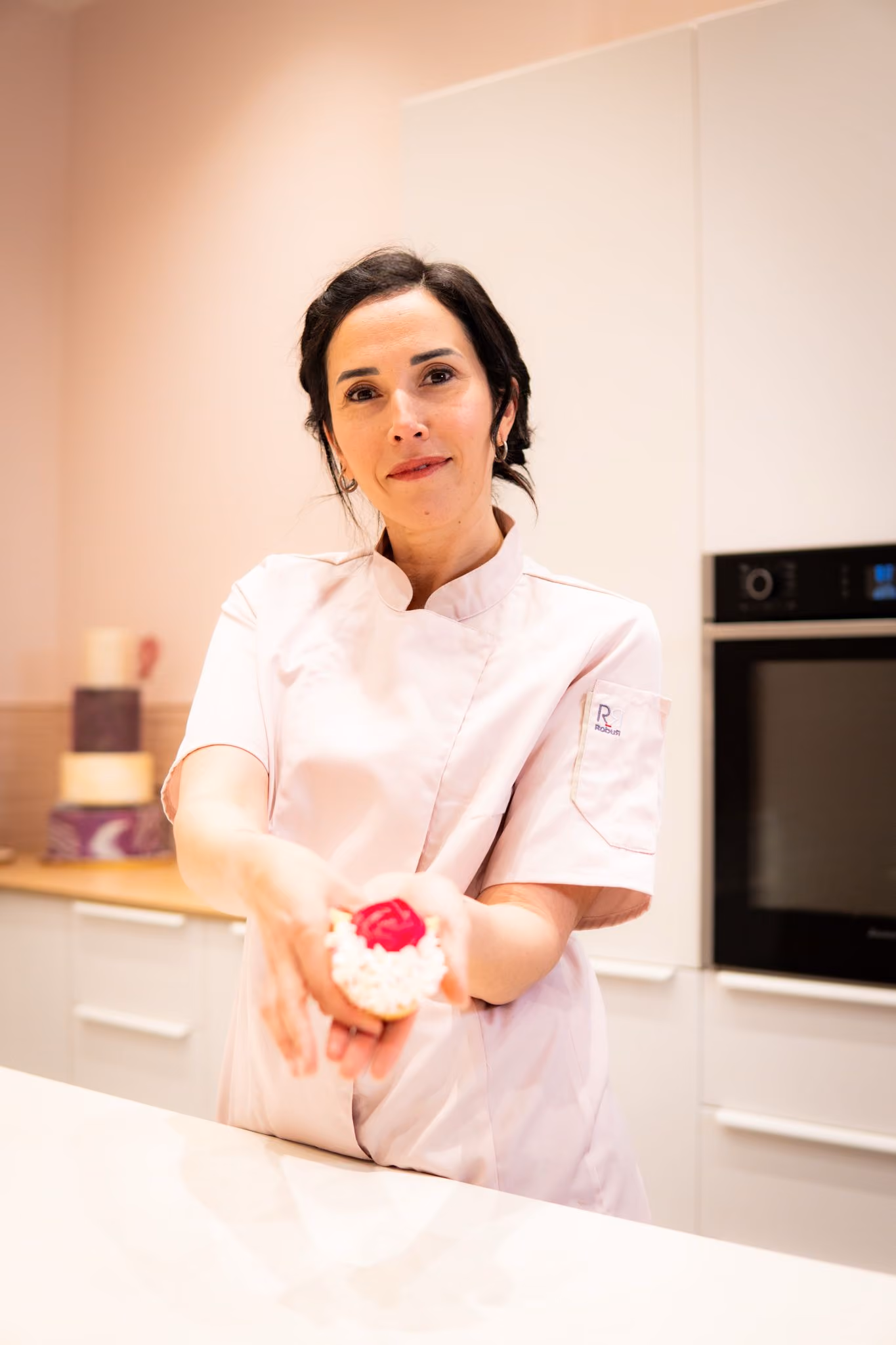 Woman in a white chef coat holding out a decorated pastry with red topping in a kitchen.