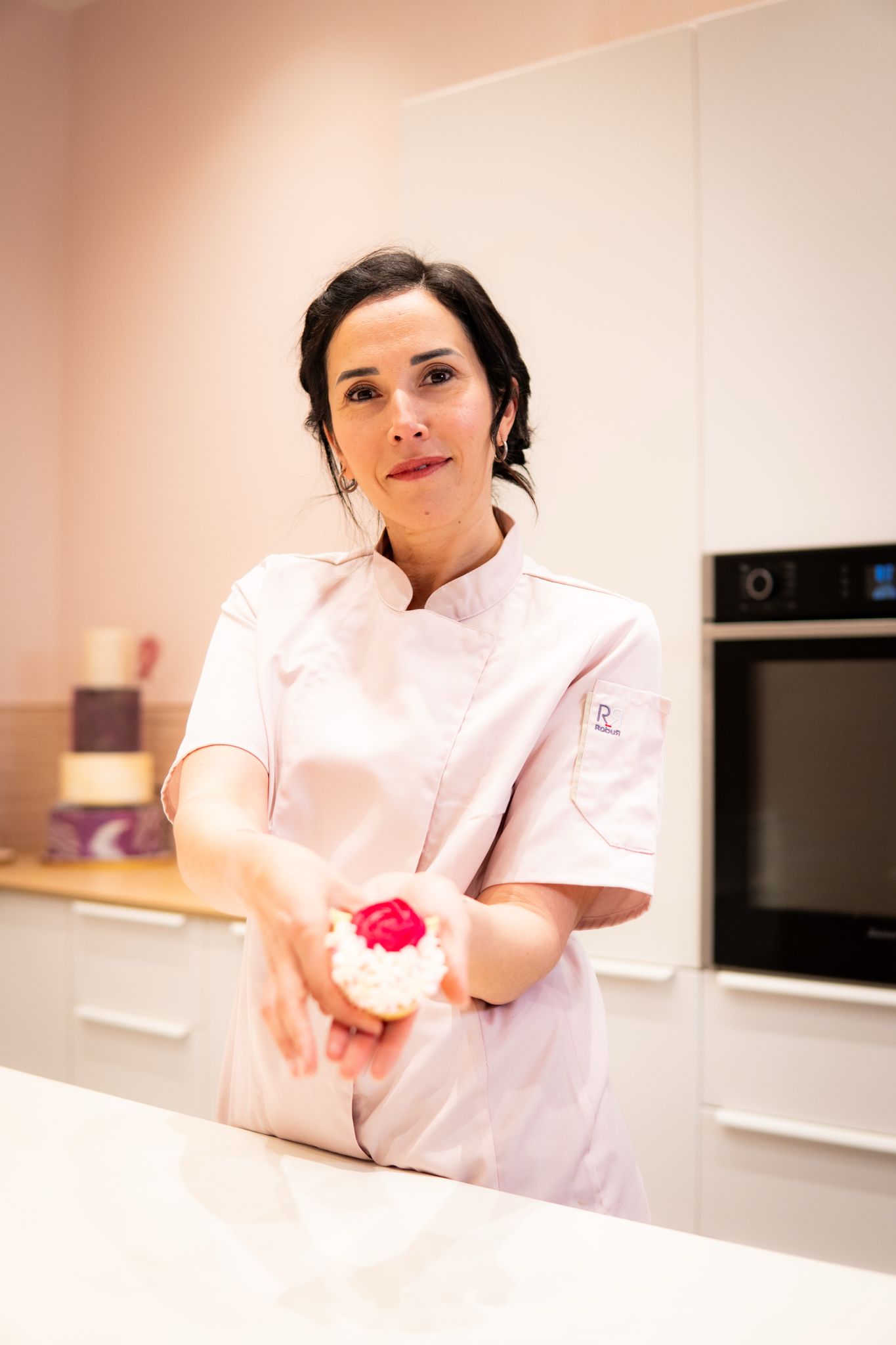 Woman in a white chef coat holding out a decorated pastry with red topping in a kitchen.