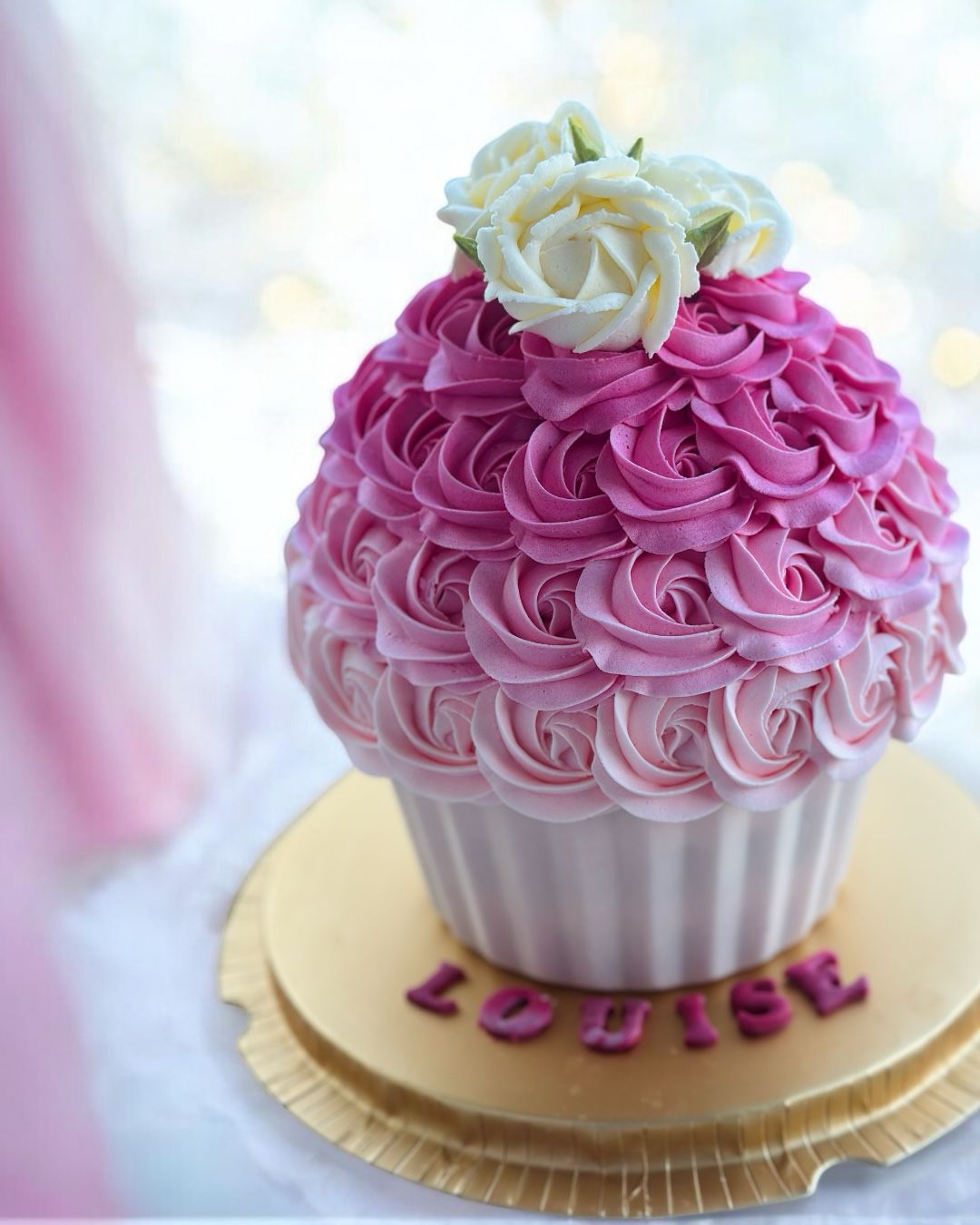 Large cupcake decorated with peach rose-shaped frosting and pearl accents, inside a clear cylindrical box with a white lid and ribbon, surrounded by purple flowers.