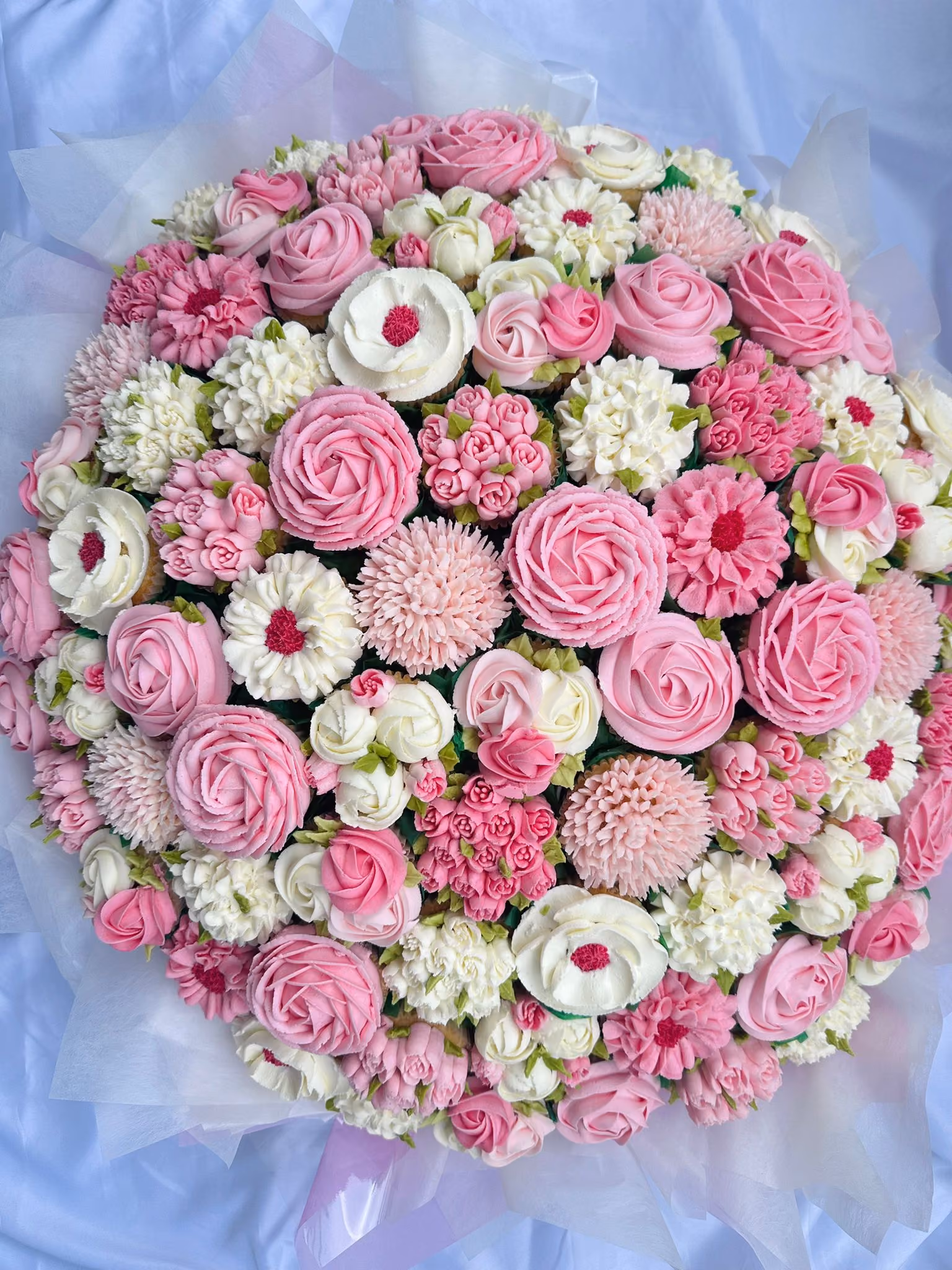 White box containing a dozen cupcakes decorated with intricate buttercream flowers in shades of red, pink, and white with green leaves.
